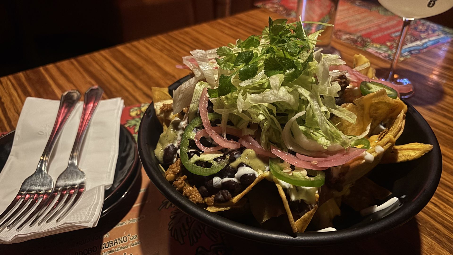 A large nacho bowl piled with shredded lettuce, cilantro on top, pickled red onions, sliced jalapeños, beans, melted cheese, sour cream, and tortilla chips around the rim on a wooden table.