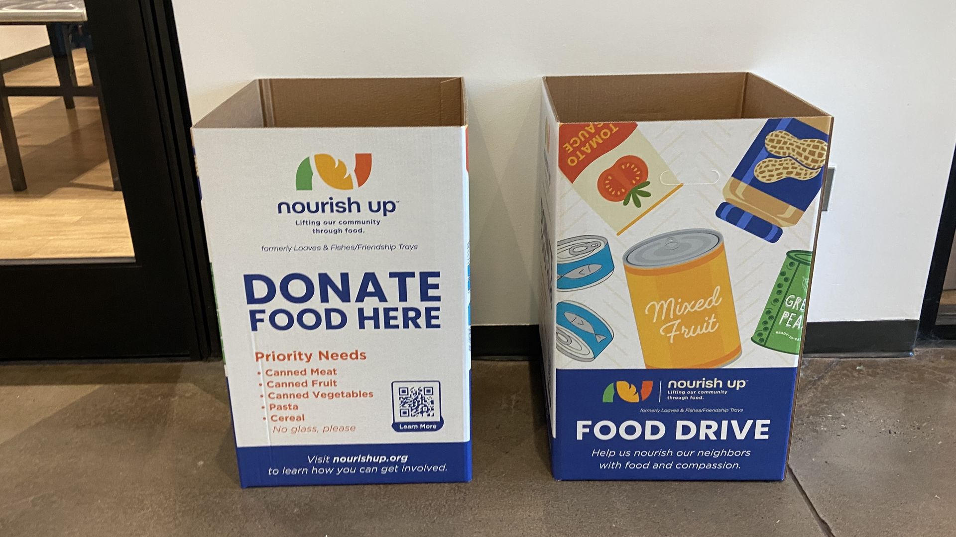 Two empty Nourish Up food donation boxes side by side, one labeled "Donate Food Here" with priority needs listed, the other decorated with canned food images and "Food Drive" text.