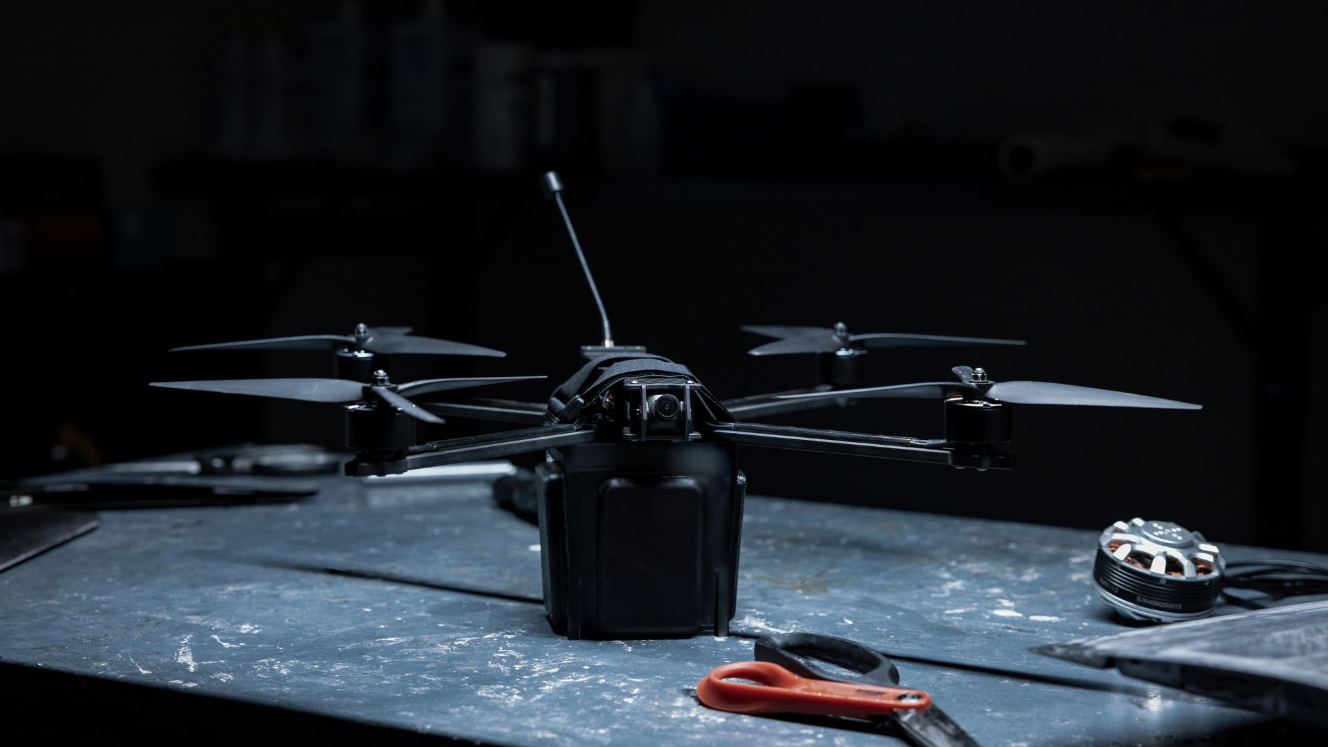 Black quadcopter drone with camera and antenna on a worn blue workbench, red scissors nearby, and a drone motor in the background under dim lighting.