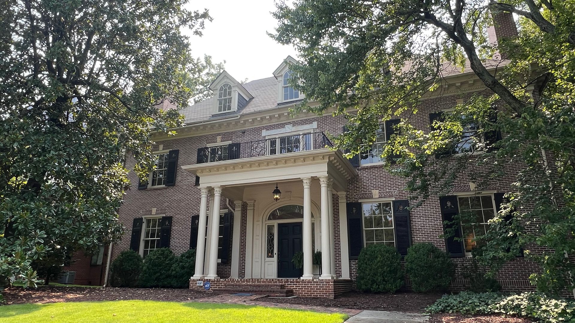 Brick two-story house with a white portico, tall columns, dormer windows, and black shutters. Surrounded by leafy trees and a manicured lawn on a sunny day.