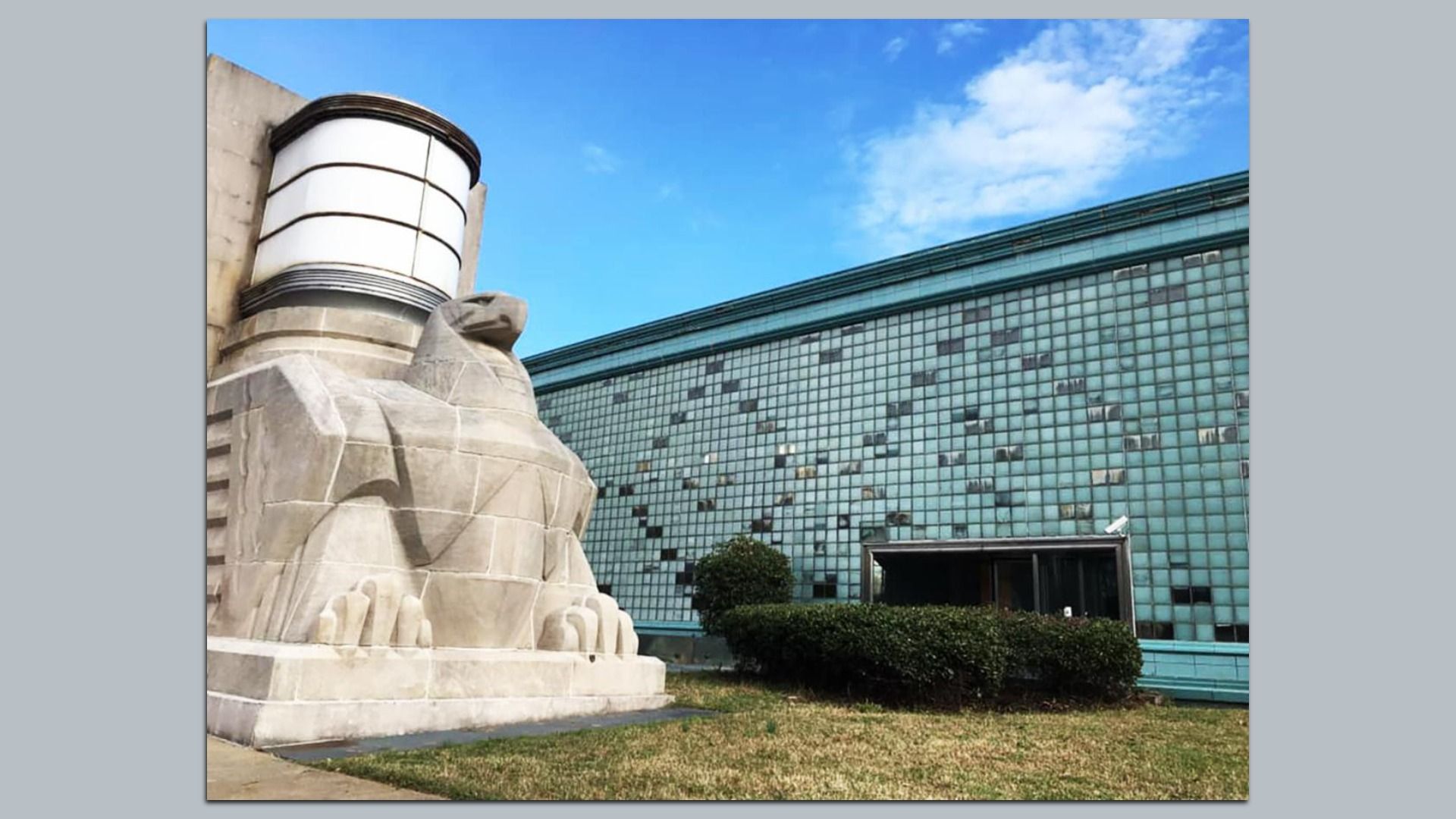 Large stone Sphinx statue in the foreground beside a turquoise glass-brick modern building under a bright blue sky, with trimmed bushes near an open entrance.