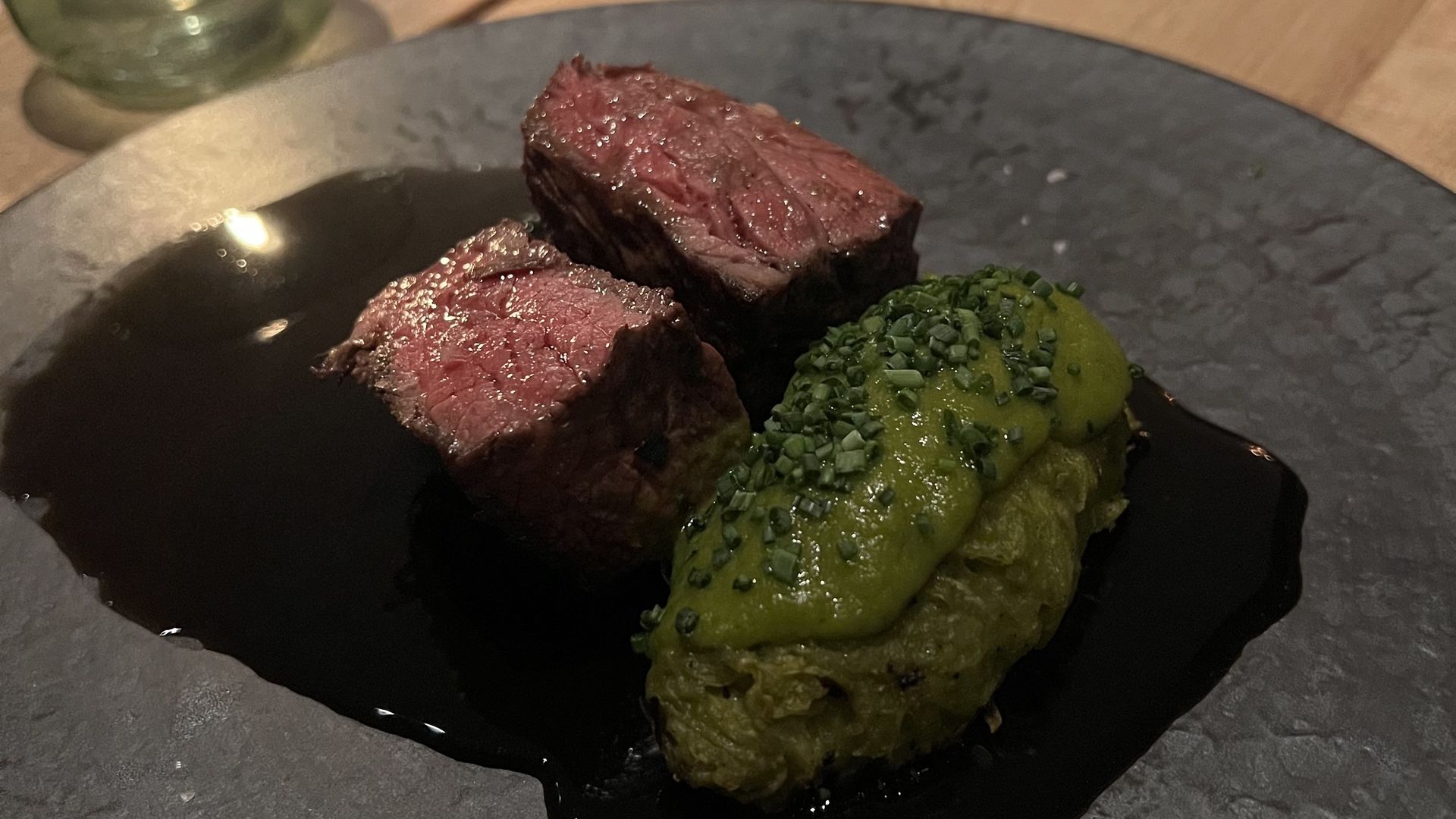 Two pieces of steak with a green herb-topped side on a black plate with dark sauce.