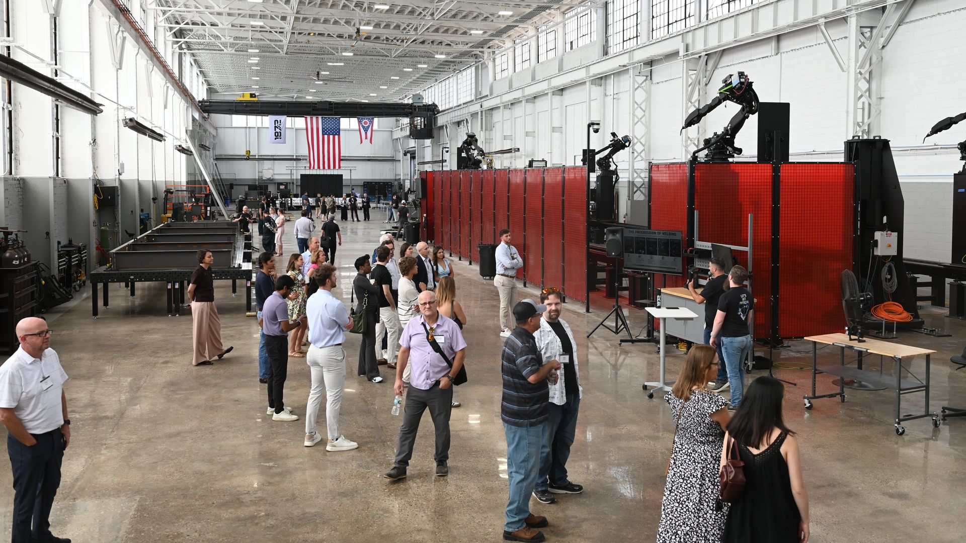 Industrial exhibition hall with white steel trusses, windows, and a crane. A crowd walks among booths near red welding barriers; robotic arms on the right; U.S. and Ohio flags in background.