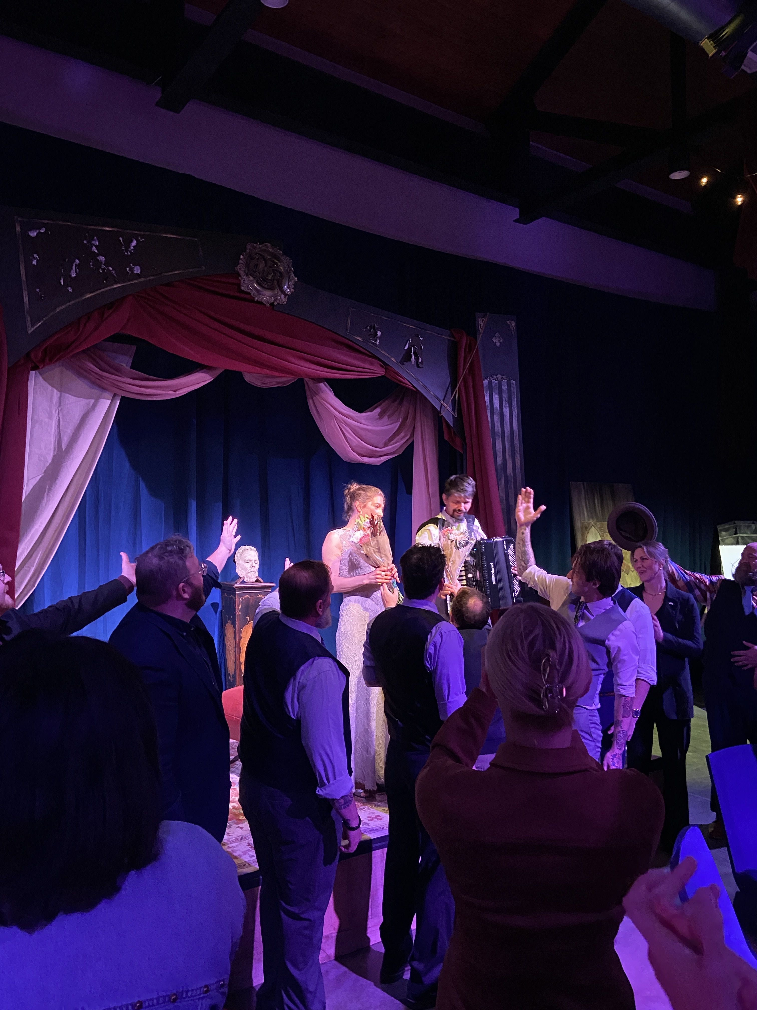 Actors on stage receiving bouquets with audience applauding; stage decorated with red and pink drapes and a bust on a pedestal under purple lighting.