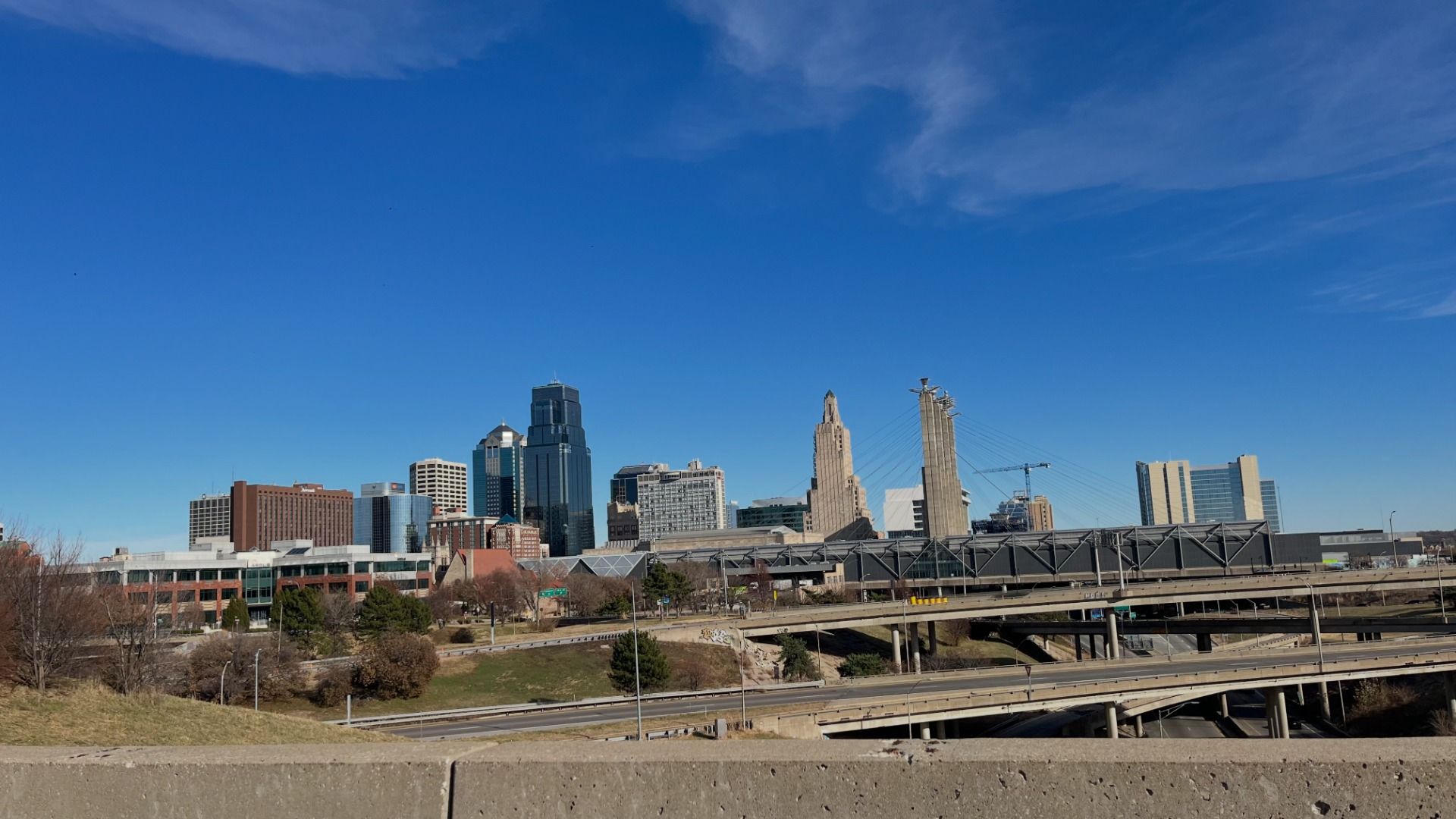 City skyline under clear blue sky with tall modern buildings, a suspension bridge, and layered highway overpasses in foreground.
