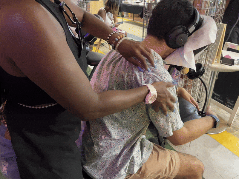 Person in floral shirt and headphones sitting in a massage chair receiving a shoulder massage from another person wearing a black sleeveless top and pink bracelet indoors with people in background.