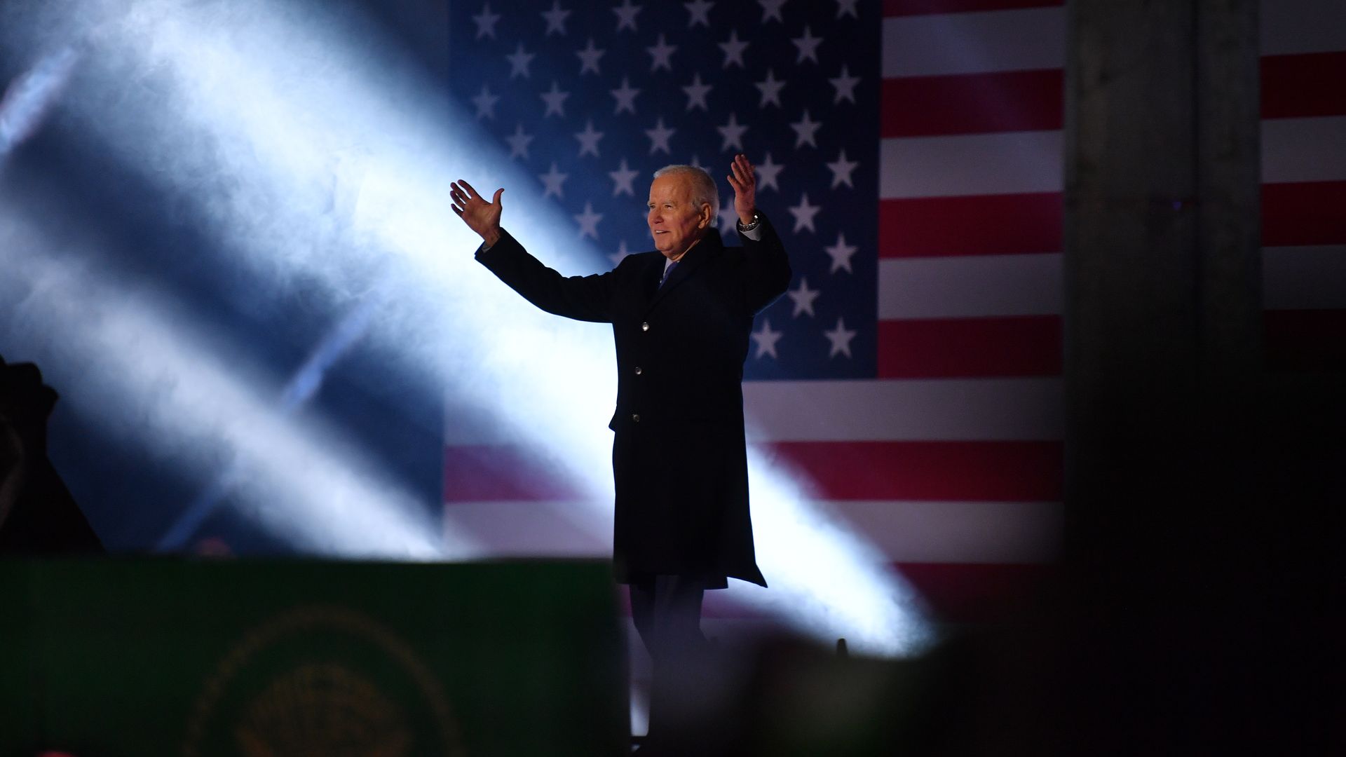 President Biden, wearing a black overcoat, stands with his arms raised in front of an American flag and spotlights shooting up.
