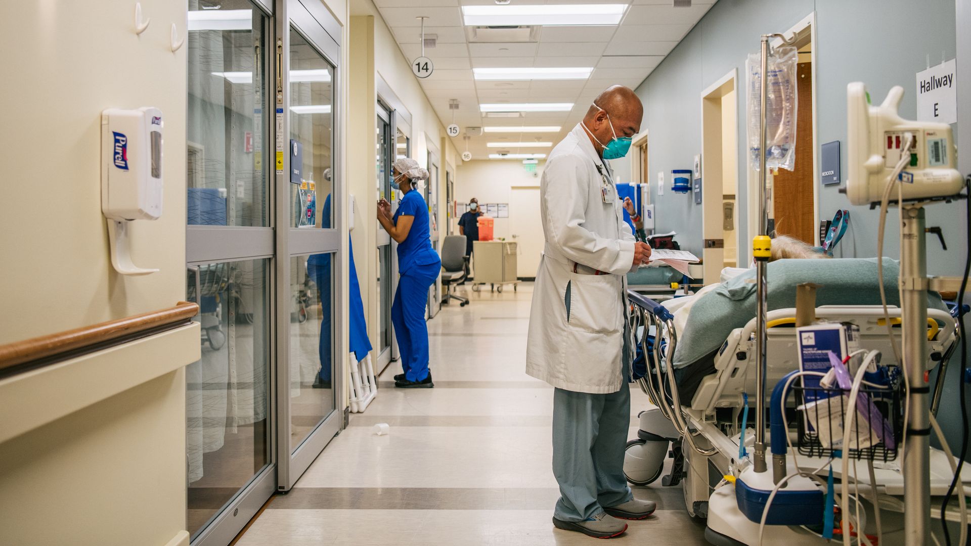 A doctor in a white coat and green mask tends to a COVID patient in a bed in a hospital hallway.