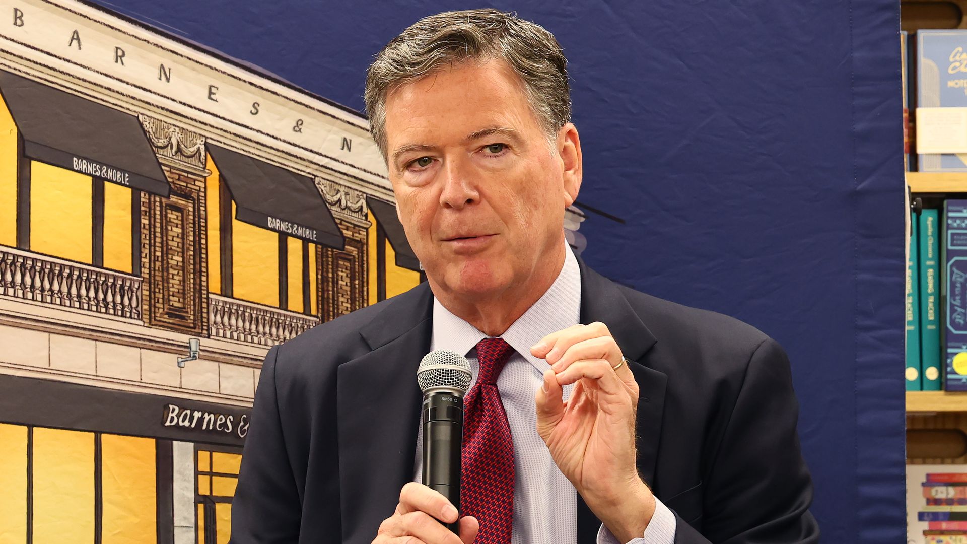 A graying, black-haired James Comey, former FBI director, gestures with his left hand as he holds a mic with his right in front of a backdrop of a building with the words "Barnes & Noble on it" to the left and a shelf full of books to the right.