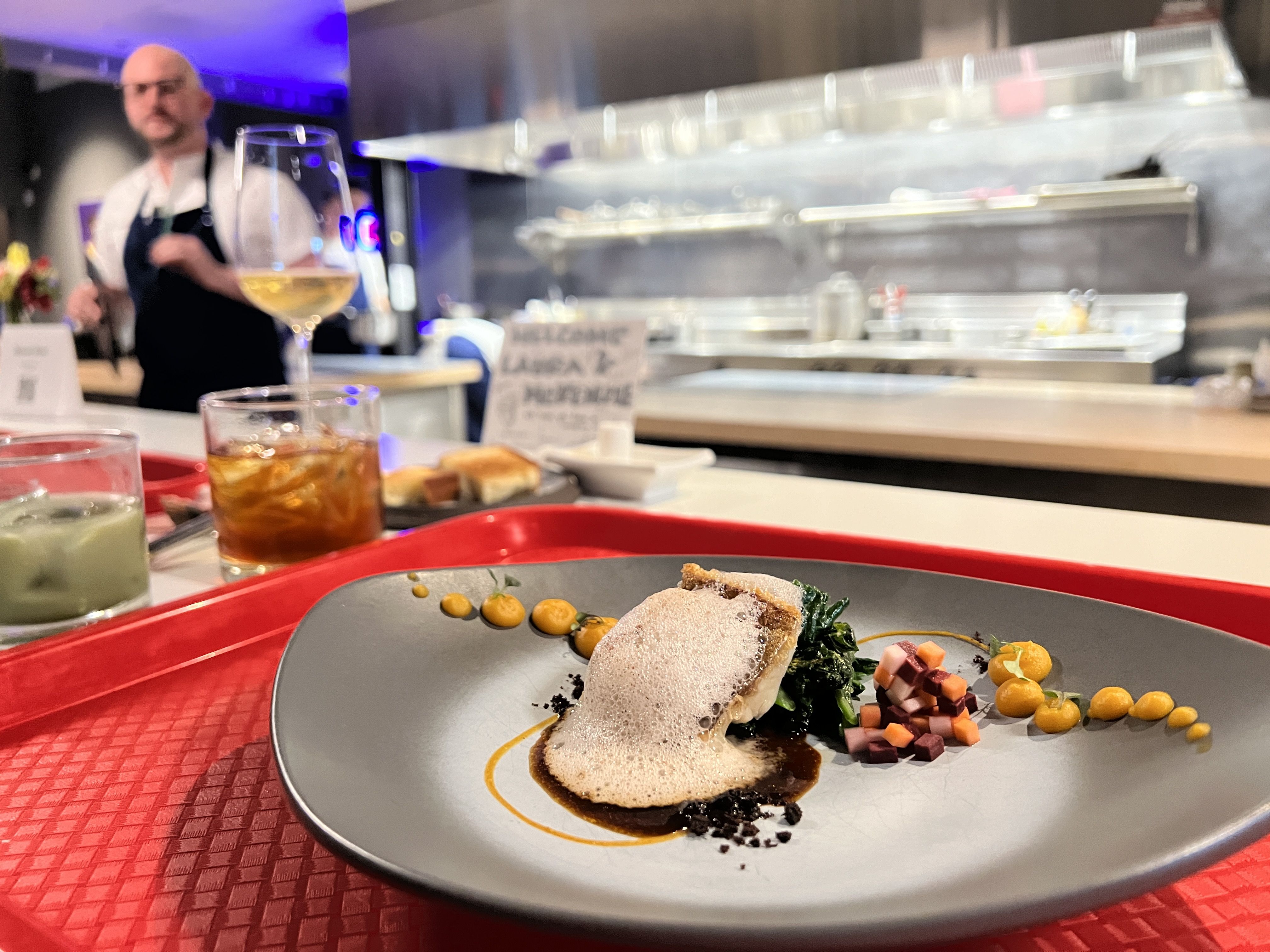 Close-up plate of fine dining with foamed sauce, greens, and diced vegetables on a gray plate with yellow dollops; blurred chef in a white shirt and stainless kitchen in the background.