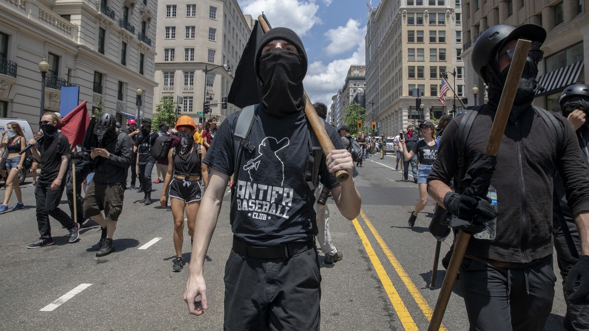 This image shows two black-clad antifa members carrying black flags down the street with their faces covered. other members follow behind them.