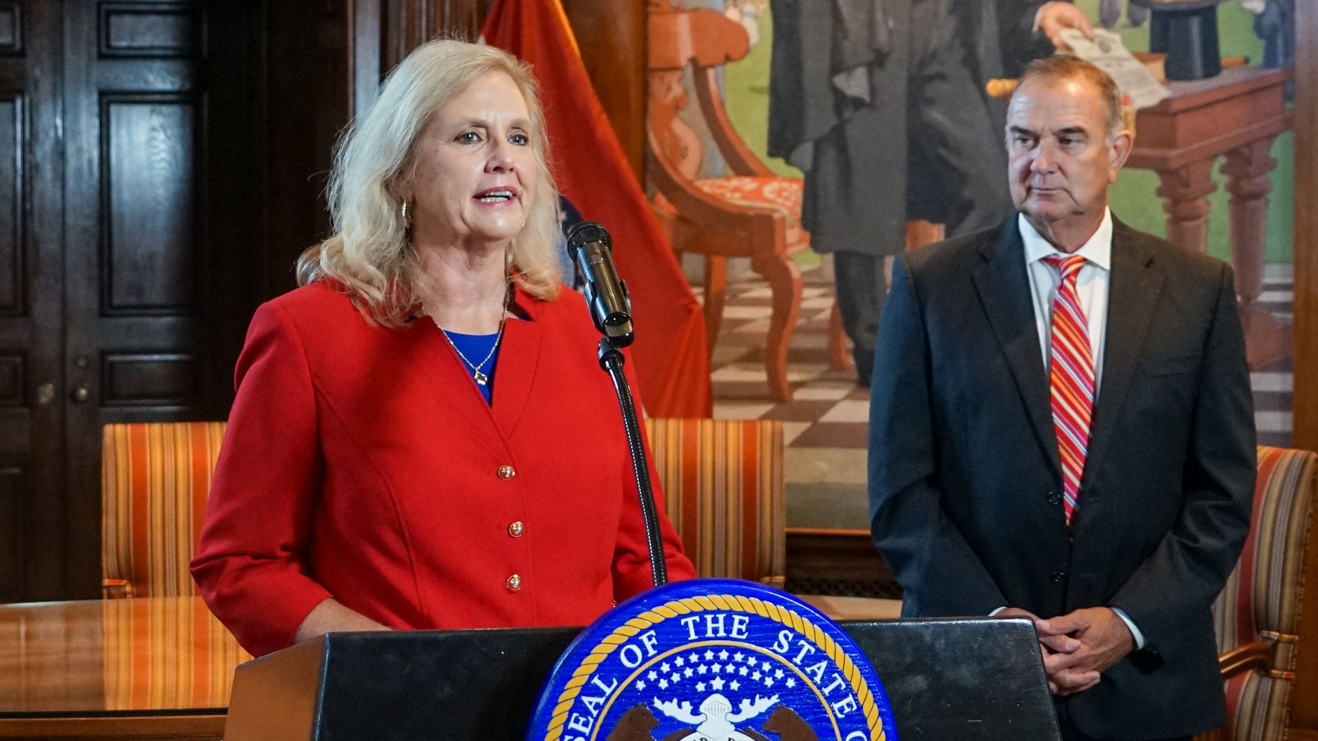 Catherine L. Hanaway in red blazer speaking at podium with Great Seal of the State of Missouri, Gov. Mike Kehoe in dark suit with red striped tie standing beside her, colorful mural and flag in background.