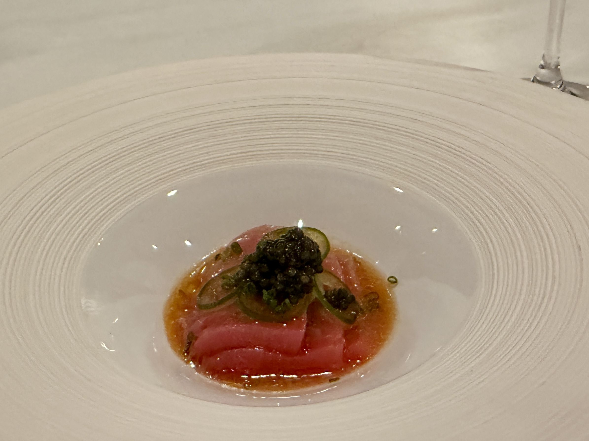 White textured bowl with a small, elegant serving of thinly sliced raw pink fish topped with dark caviar and thin green slices in a light orange sauce.