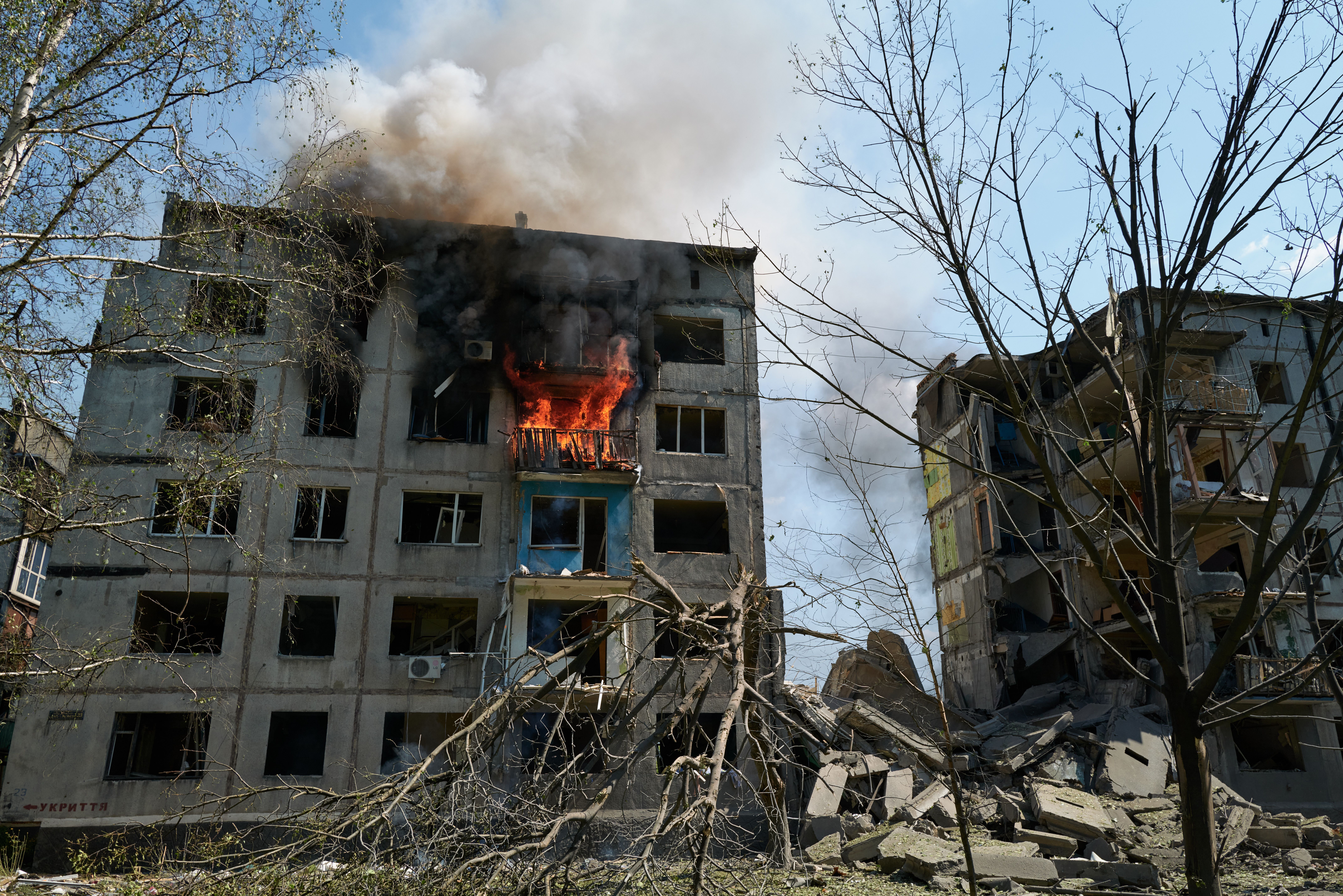 A damaged multi-story building with a balcony engulfed in flames and thick black smoke, surrounded by debris and leafless trees under a clear sky.