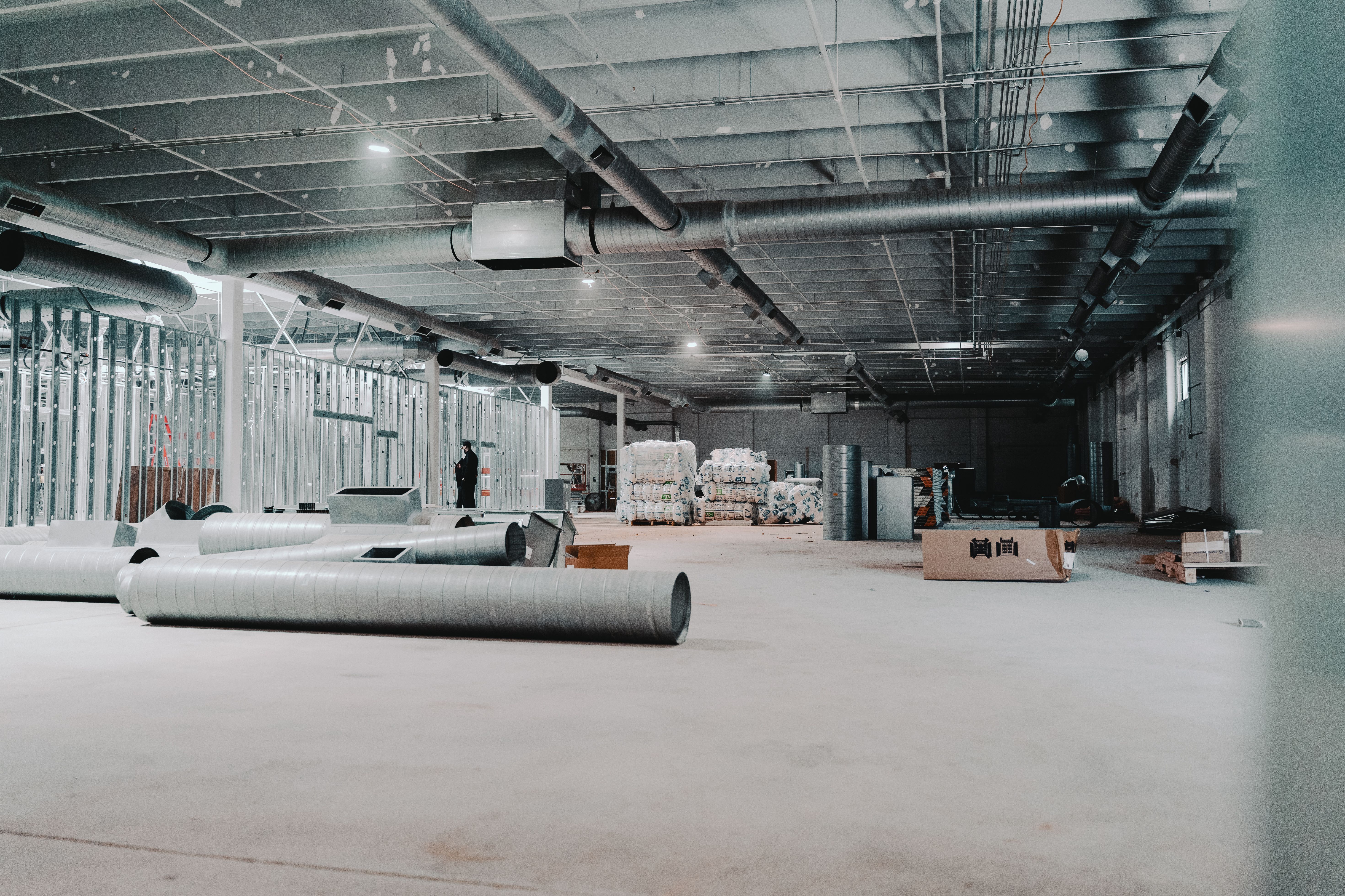 Industrial construction site with exposed silver ducts and pipes overhead, metal framing along the left, stacks of pallets and boxes, and a worker standing near the back.