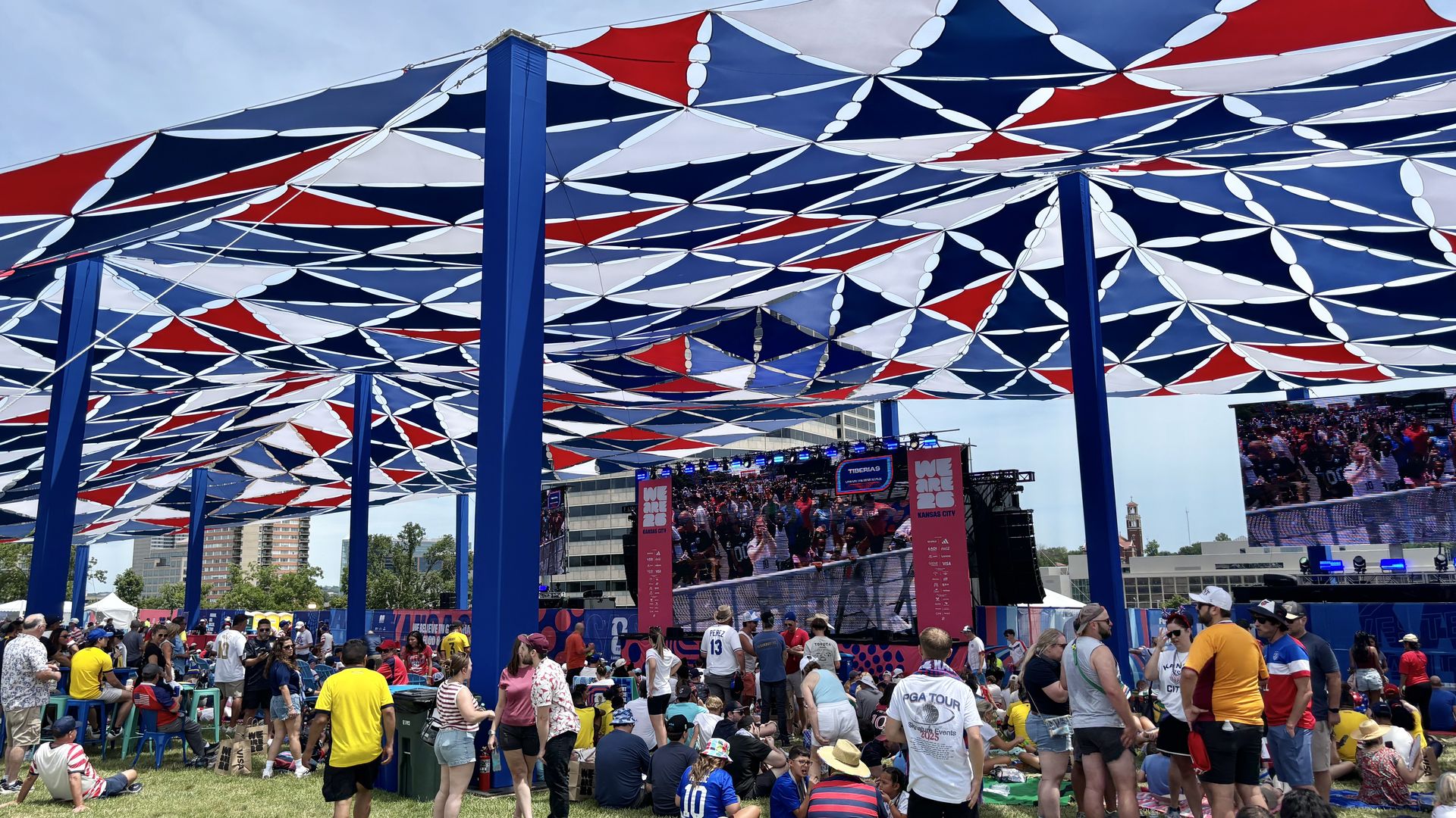 Crowd at an outdoor event beneath a red, white, and blue geometric fabric canopy supported by tall blue pillars; a stage with large video screens visible in the background.