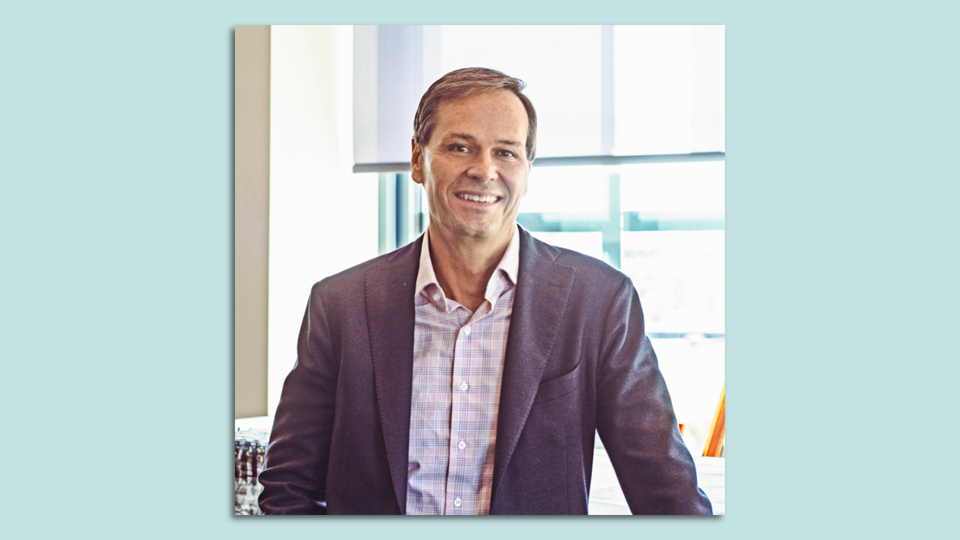 A man in a blue blazer stands in front of window with a blue background