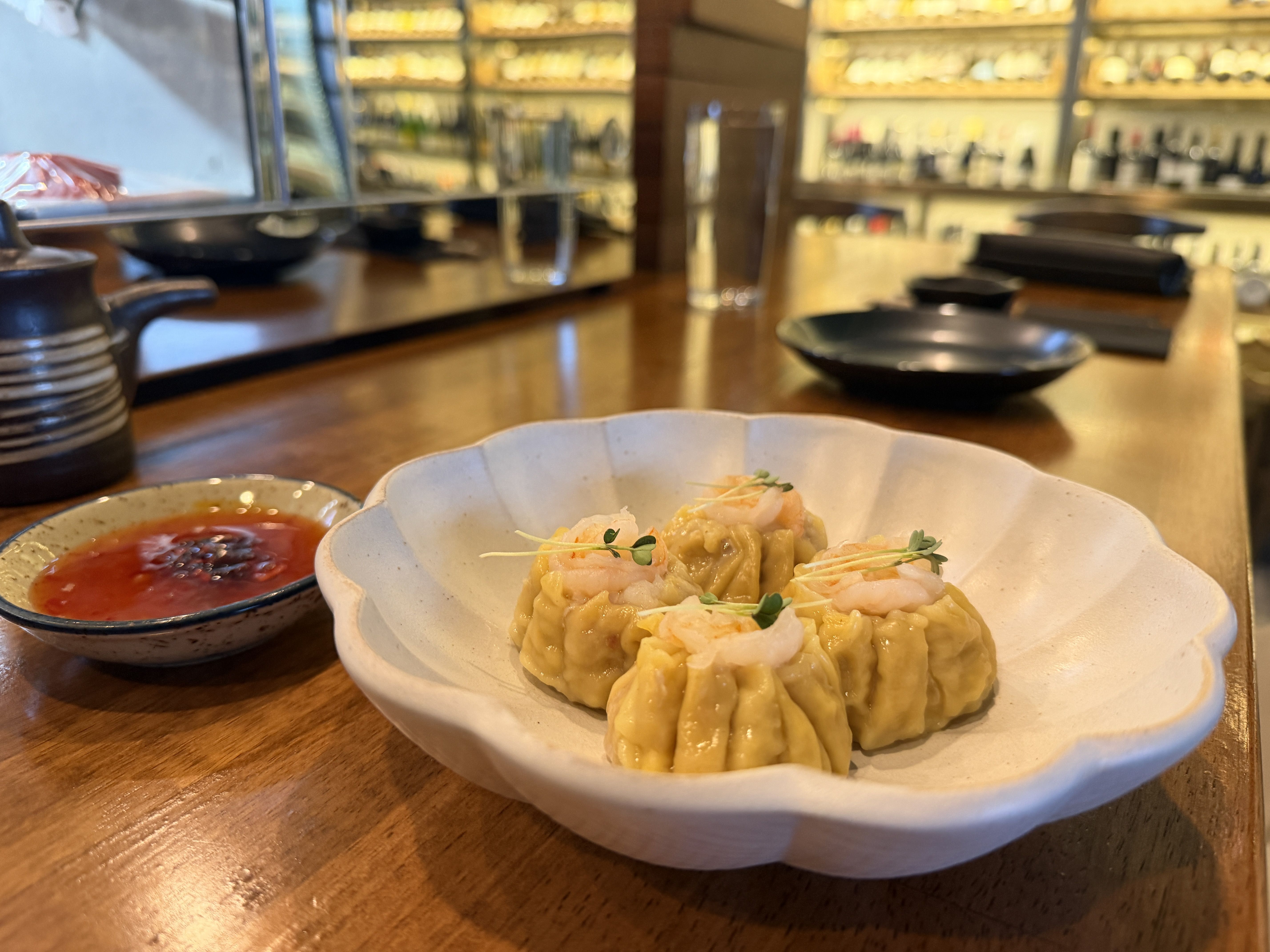 Four shrimp-topped dumplings in a white scalloped bowl with a small dish of red dipping sauce on a wooden table in a warmly lit restaurant setting.