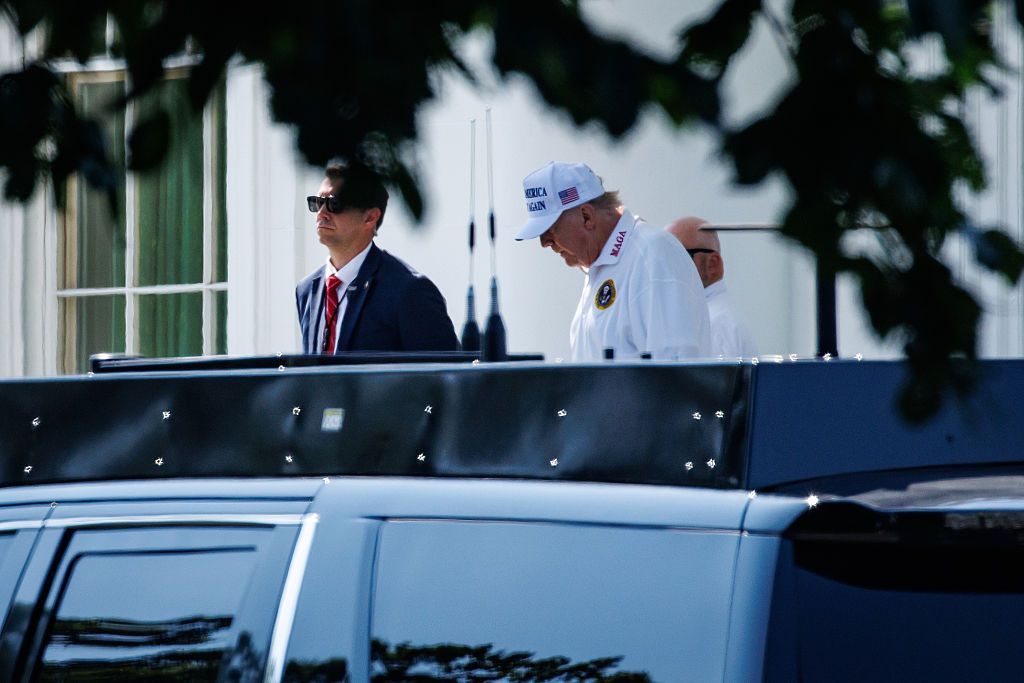 A photo showing President Trump wearing an all-white outfit. MAGA is printed on his collar and he's wearing a Make America Great Again baseball cap. A man wearing sunglasses and a red tie stands in the frame. They appear to be heading into a vehicle.