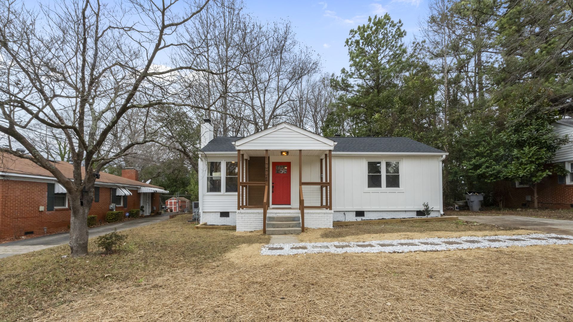 Small white house with a front porch and red door, surrounded by bare trees and brown grass, neighboring red brick houses on either side under a cloudy blue sky.