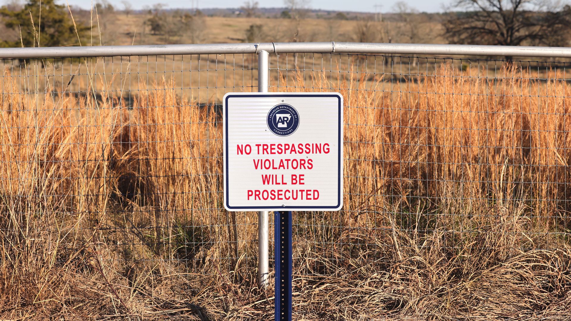 A sign reading "no trespassing violators will be prosecuted" stands before a wire fence with an empty field of dry grass in the background.