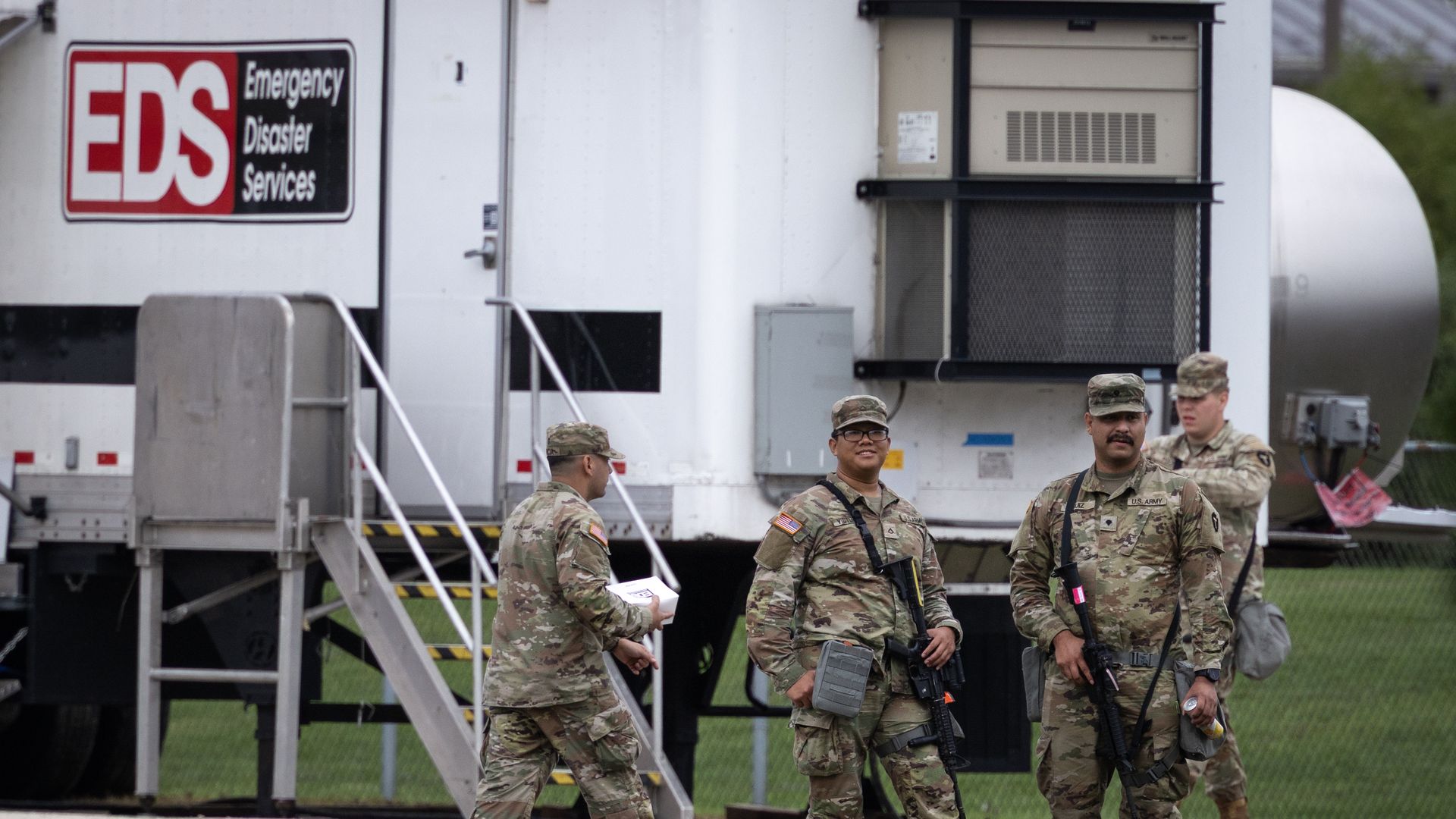 Four male members of the Texas National Guard, wearing their khaki uniform and carrying large, black guns, at the Elwood Army Reserve Training Center in Elwood, Illinois, in front of an Emergency Disaster Services sign on a truck. 