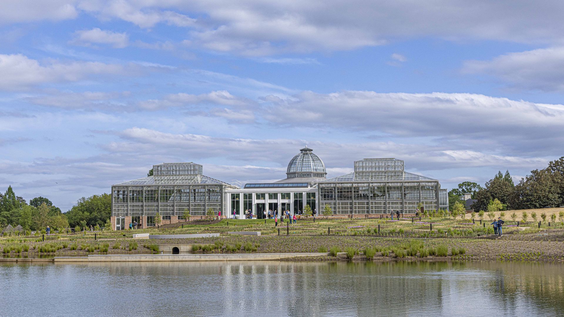 Glass conservatory with a domed center and surrounding glass wings sits by a pond. People walk along the foreground path, trees and greenery framing the scene under a blue, cloudy sky.