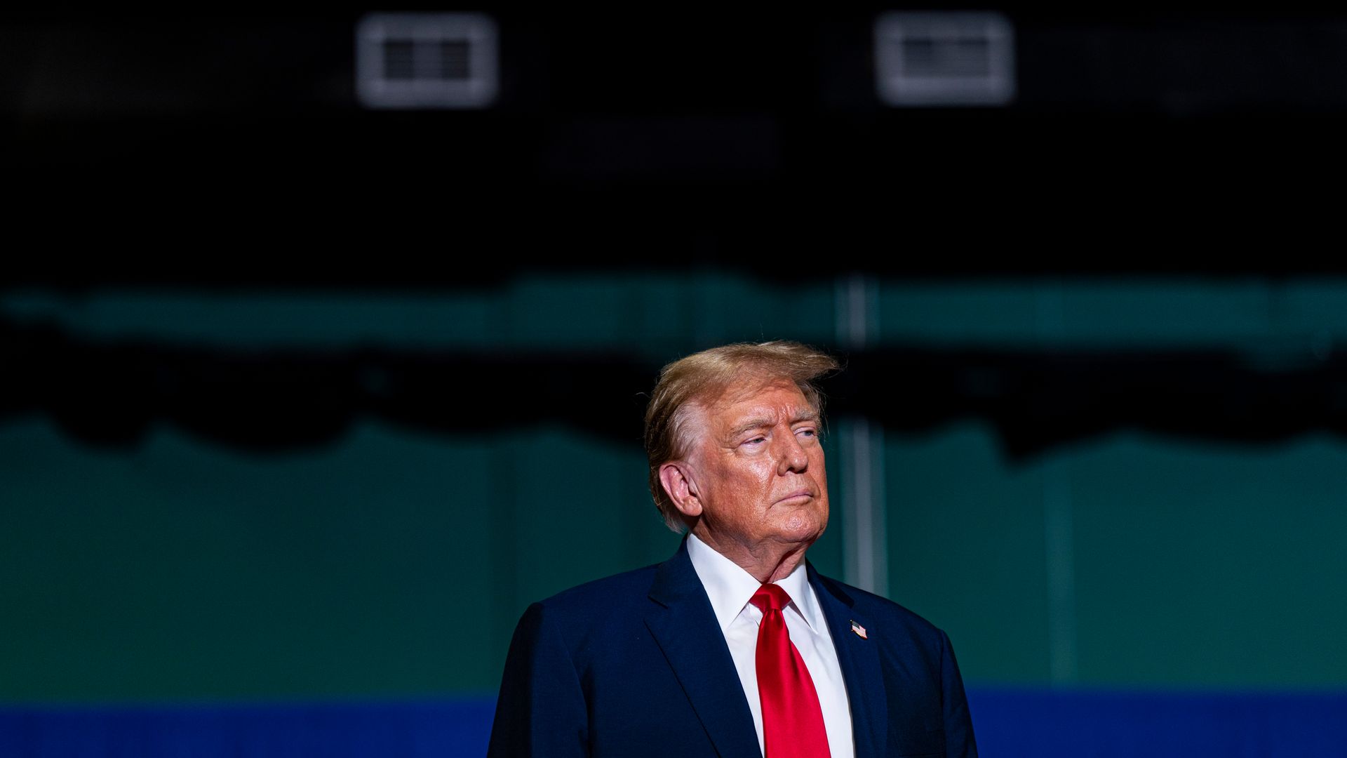 Former President Trump, wearing a blue suit jacket, white shirt and red tie, standing against a green and blue wall covered in shadows.