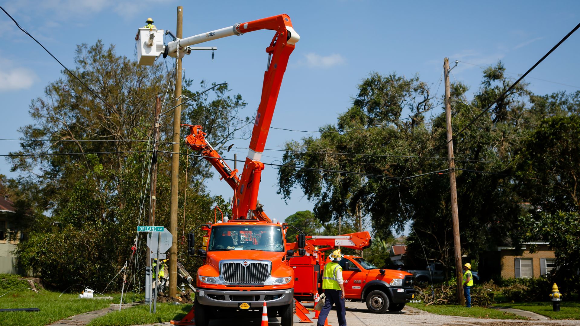 A worker tends to gear at the top of an electrical pole from inside a bucket truck, which is extended to reach the top of the pole. On the ground, several more workers view the progress.
