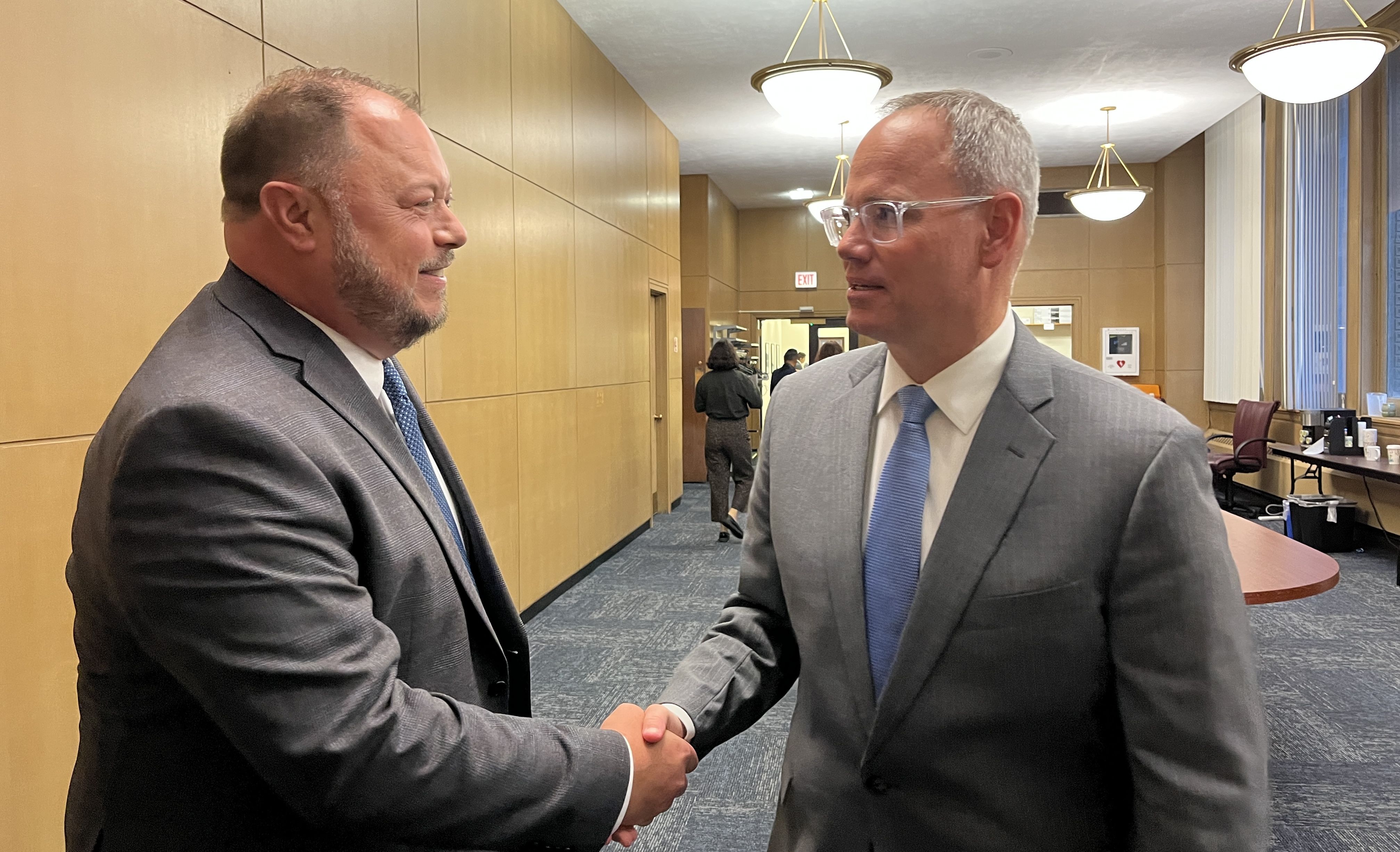 Two men in gray suits and blue ties shake hands in a wood-paneled office hallway with blue carpet and round overhead lights.