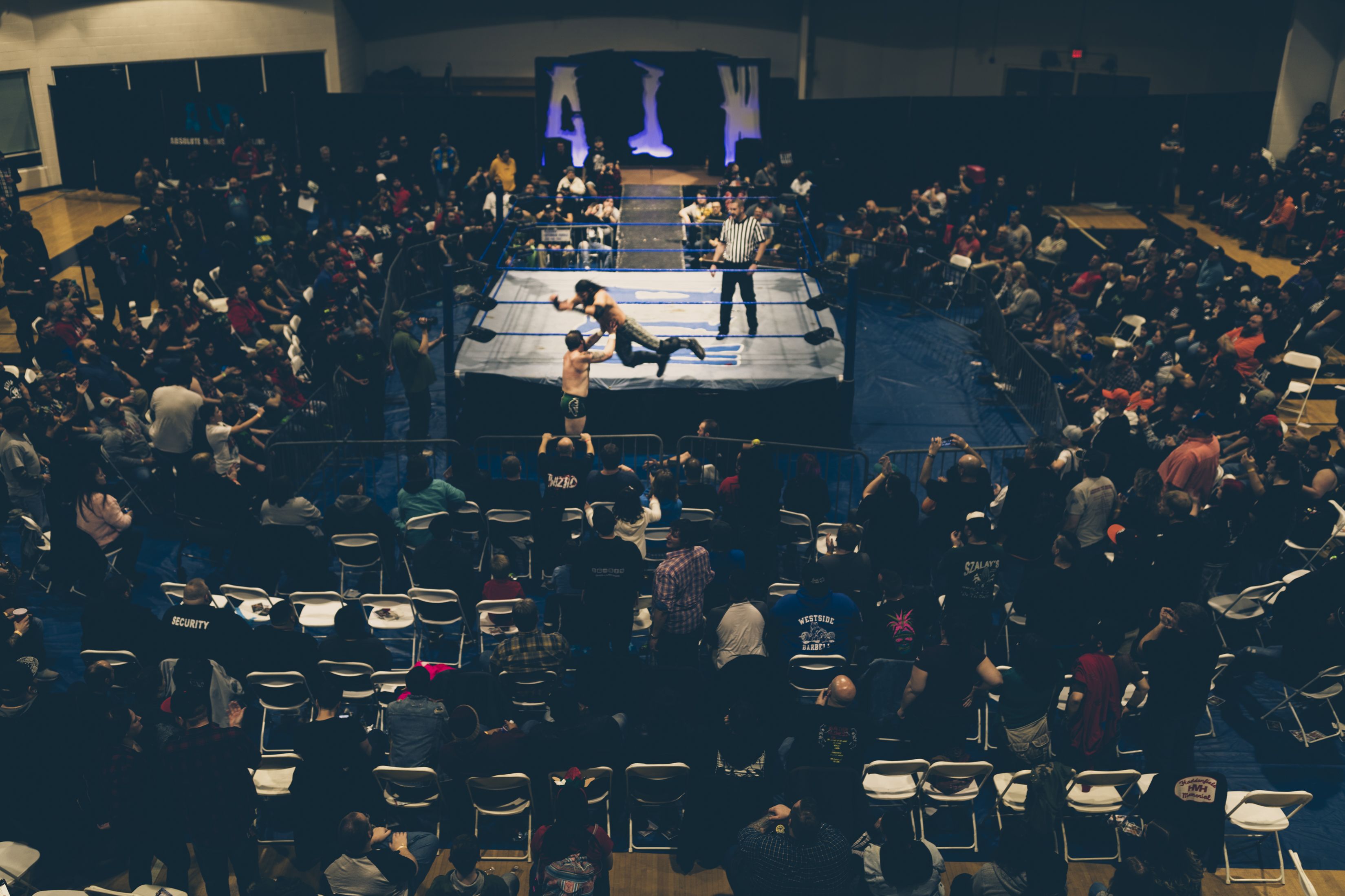 Indoor wrestling ring with blue ropes at center, surrounded by a dense crowd seated and standing. A wrestler leaps toward an opponent as the referee watches.