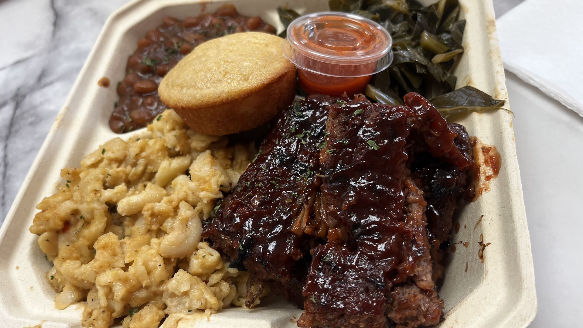 Ribs, mac & cheese, collard greens, beans and corn bread in a to-go box. 