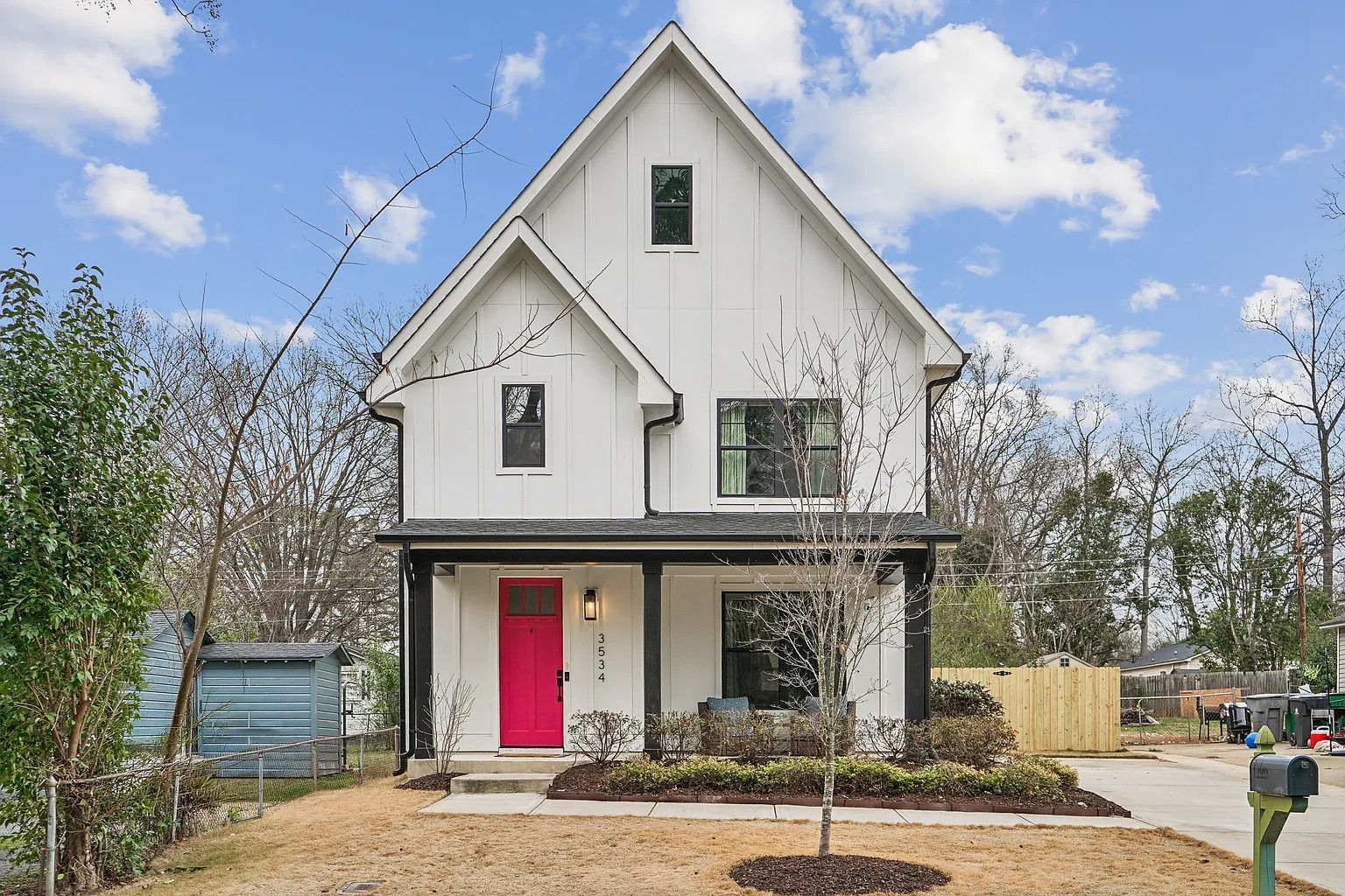 White two-story house with black trim, bright pink front door, porch with chair, small leafless tree and shrubs in front yard, beige grass, blue sky with clouds.