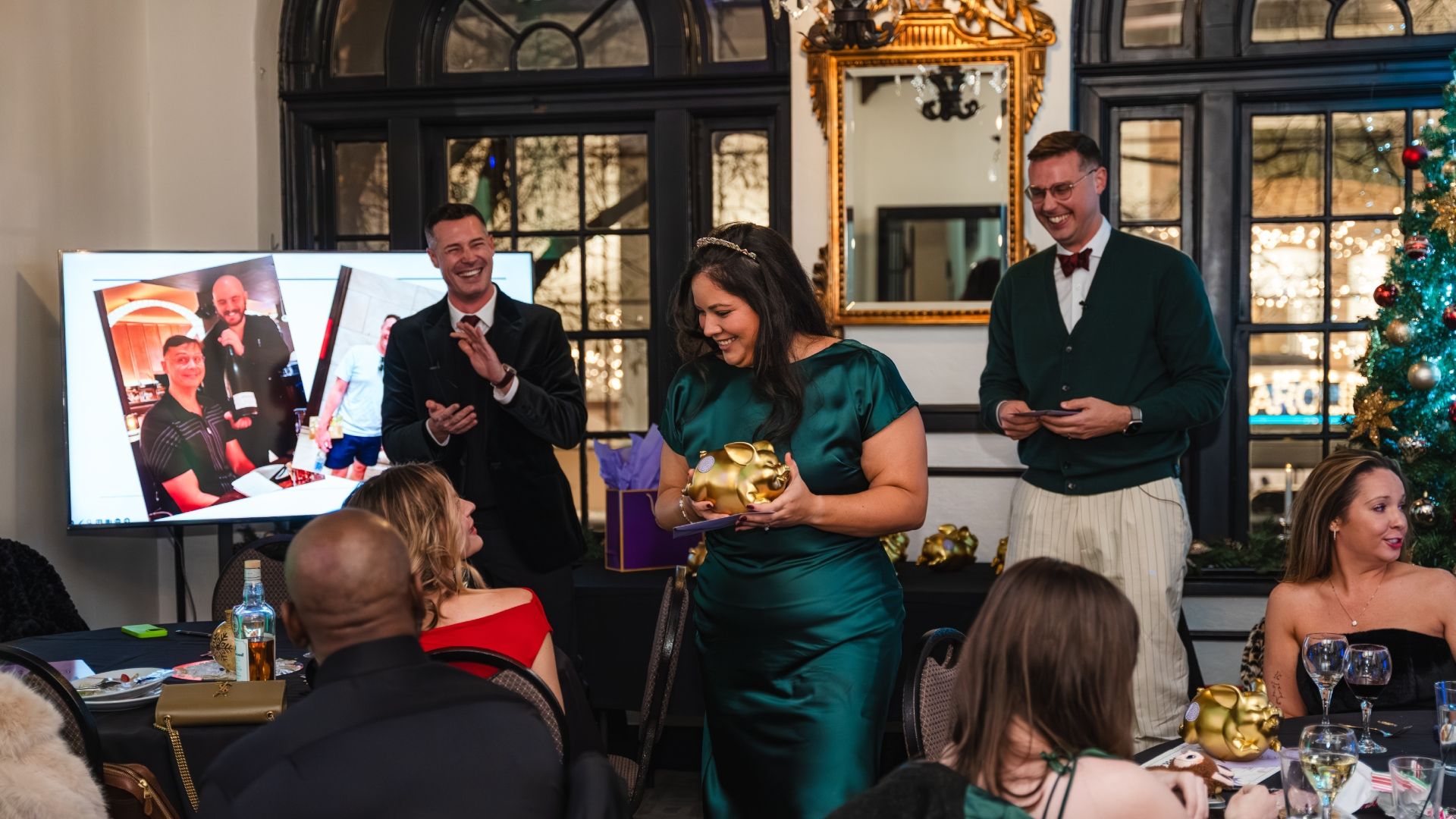 Holiday party scene: woman in emerald green holds a gold pig statue and a card, smiling; two men in suits laugh beside her. A Christmas tree and a gold-framed mirror decorate the background.