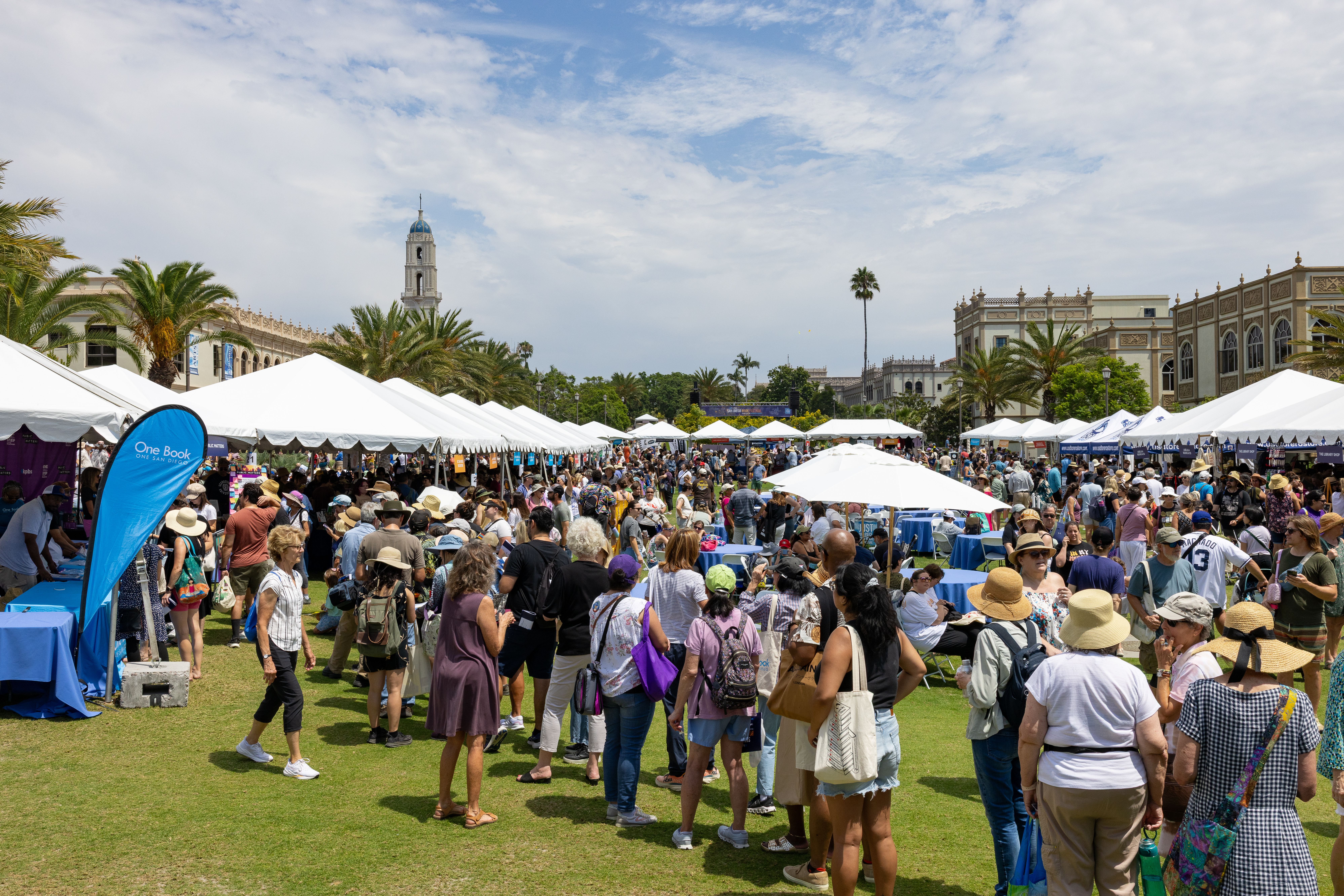 Busy outdoor market with white tents and crowds, palm trees, a distant tall clock tower and ornate buildings under a blue, partly cloudy sky.
