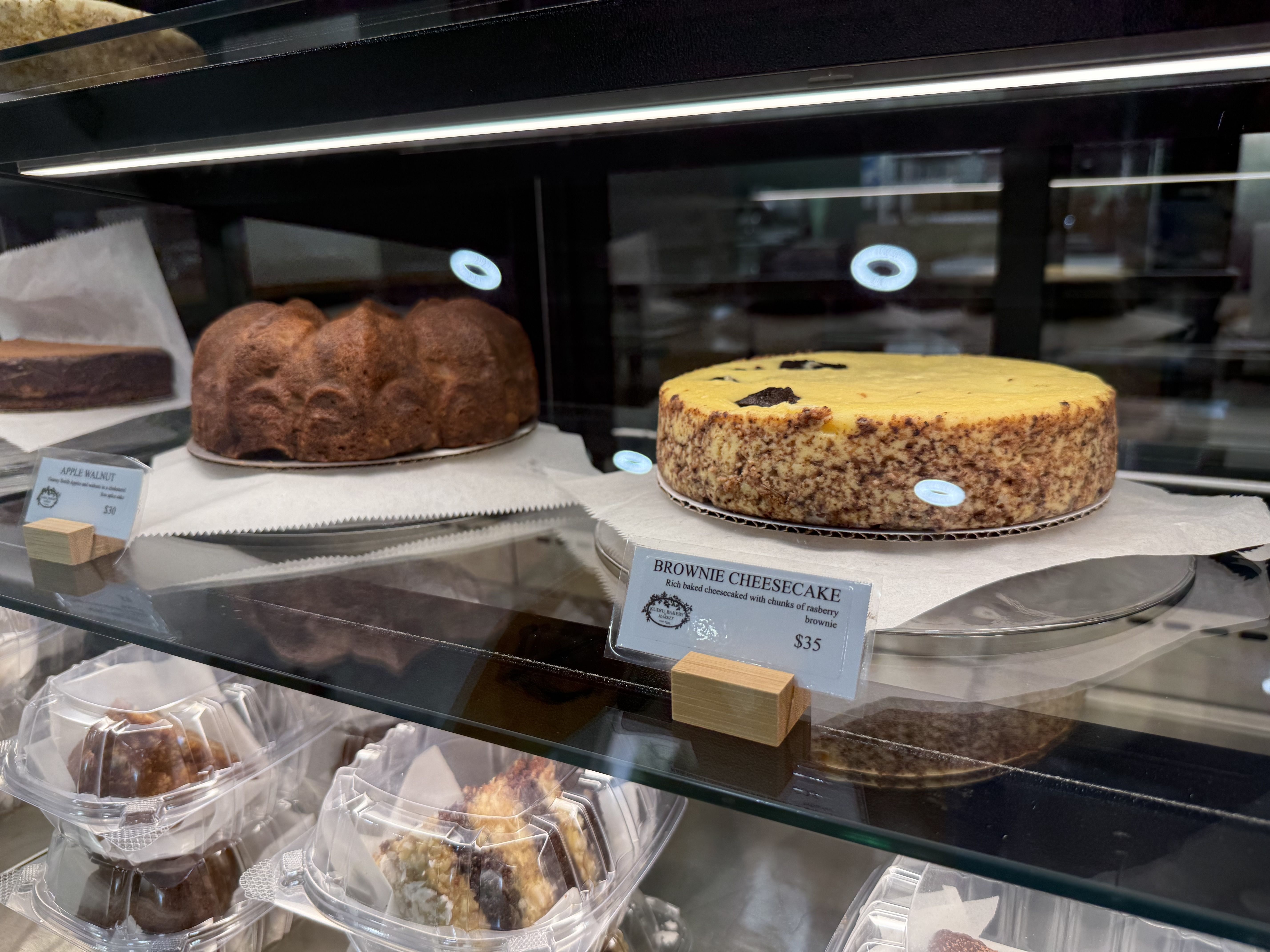 Glass display case with two cakes on white paper. Left is a brown apple walnut bundt cake, right is a round brownie cheesecake with yellow top and chocolate speckles. Price tags visible.