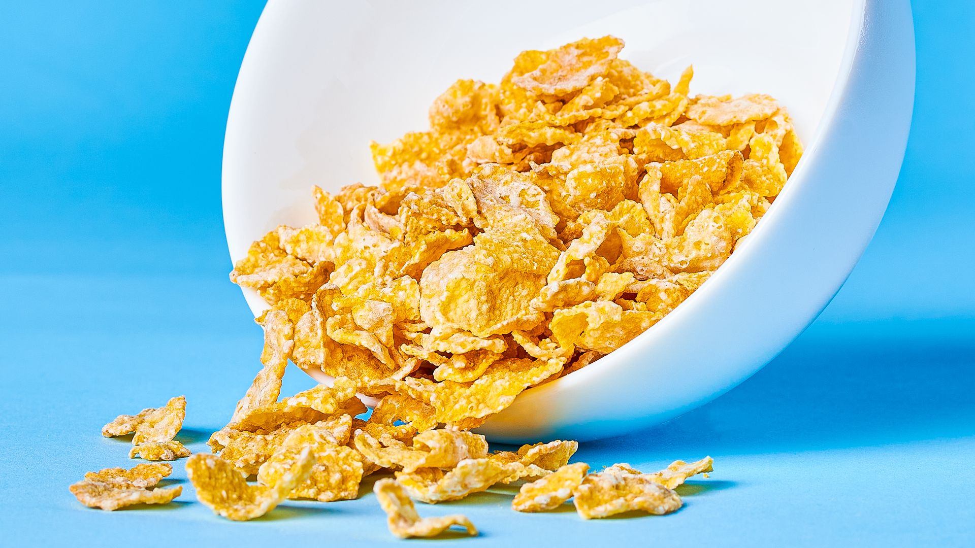 White bowl on a vivid blue backdrop, tipped to spill golden cornflakes across the surface with flakes scattered around.