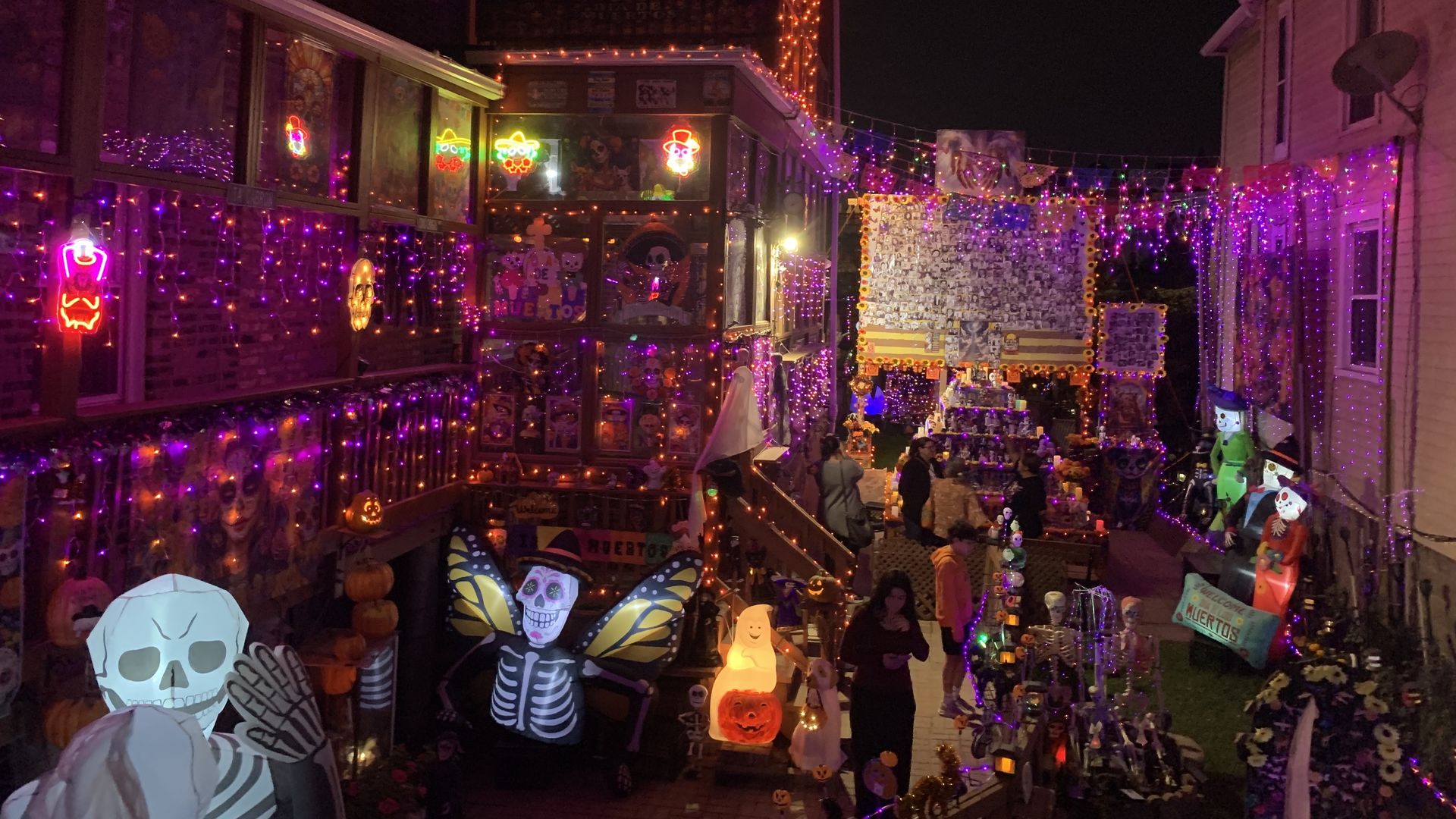 Nighttime street festival with purple and orange string lights, skull and pumpkin decorations, neon shop signs, and stalls displaying Halloween decor along a festive alley.