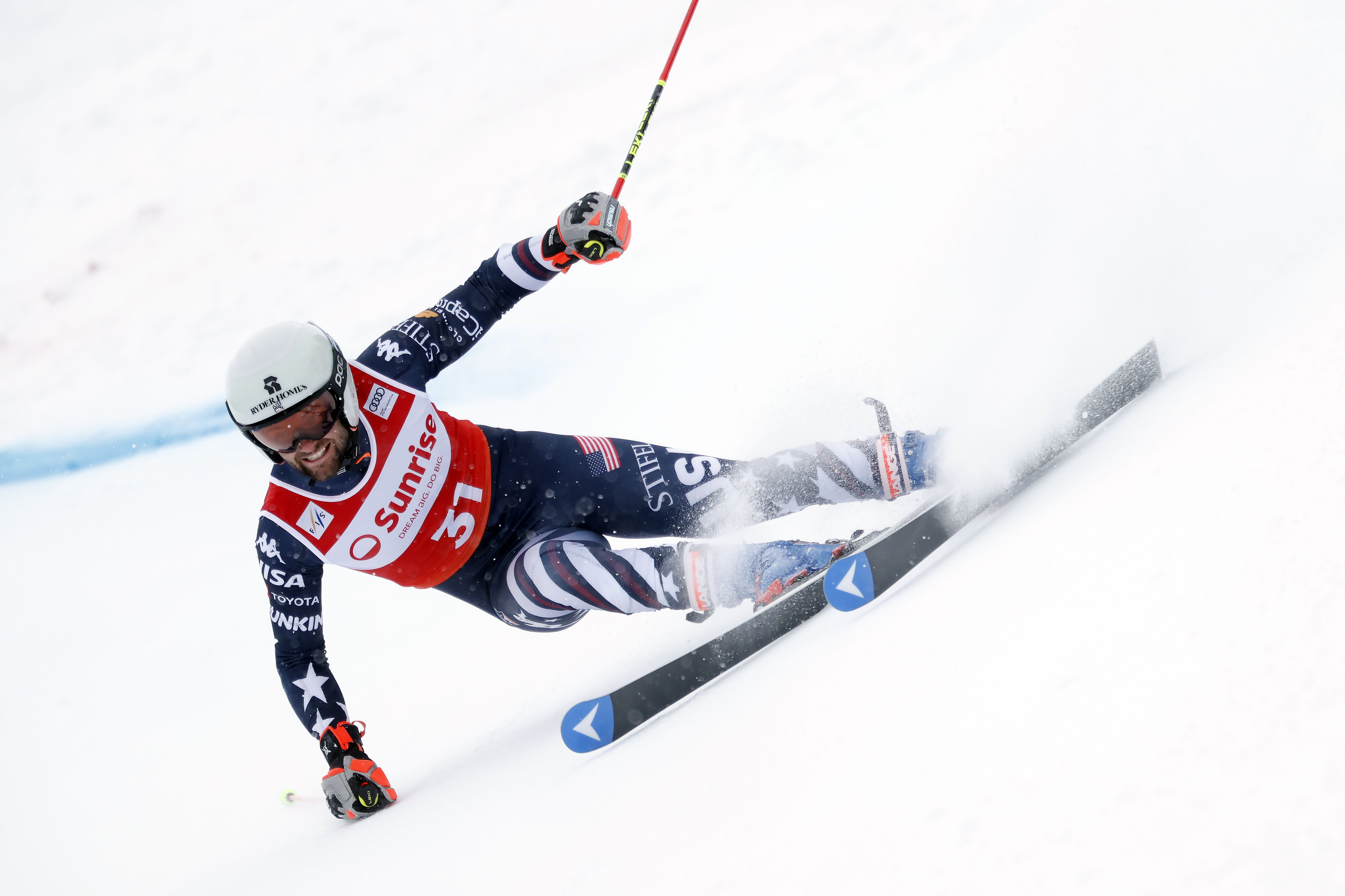Skier wearing a white helmet and blue USA racing suit with stars, red bib number 31, turning sharply on snowy slope, creating snow spray, holding red and yellow ski poles.