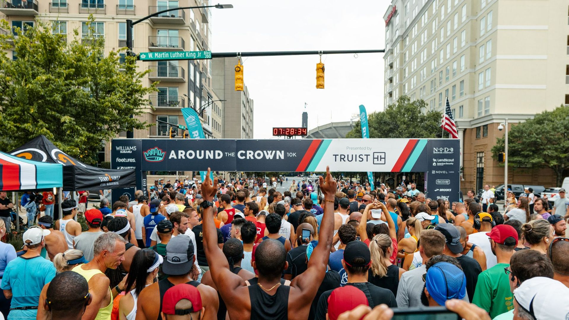 Crowd of runners gathered at the start line of the "Around the Crown 10K" race under W. Martin Luther King Jr Blvd street sign, with a timer and urban buildings in the background.