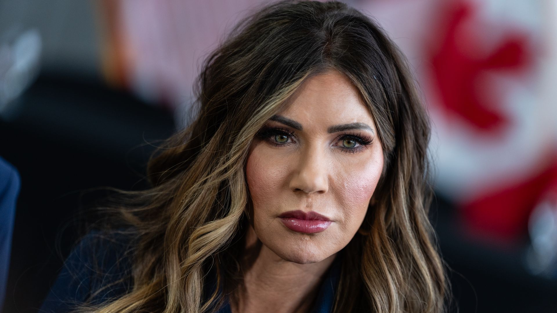 A heavily made up Homeland Security Secretary Kristi Noem, with dyed dark hair and lighter highlights, and full, pink lips pouts as she stares ahead.