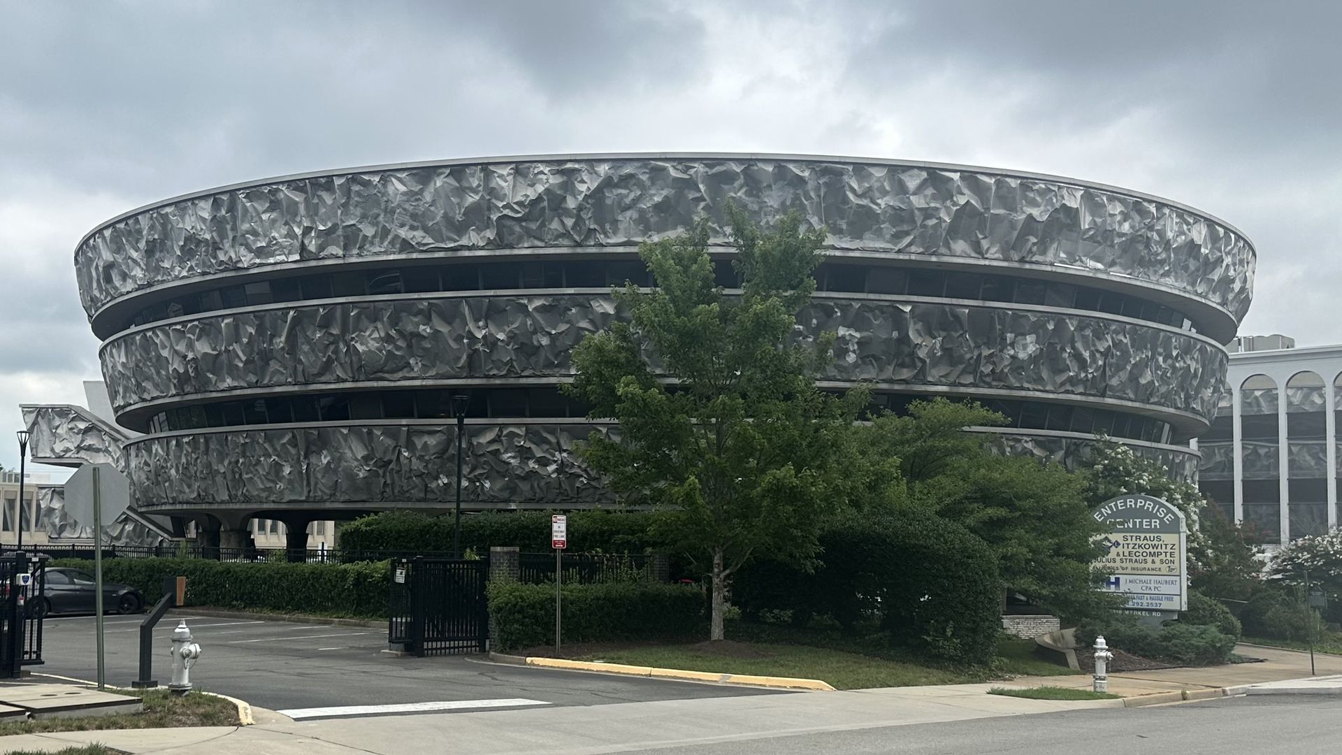 A round, multi-tiered building with a crumpled silver metal facade, set against a cloudy sky; green trees obscure the lower levels, and a nearby sign reads "Enterprise Center".