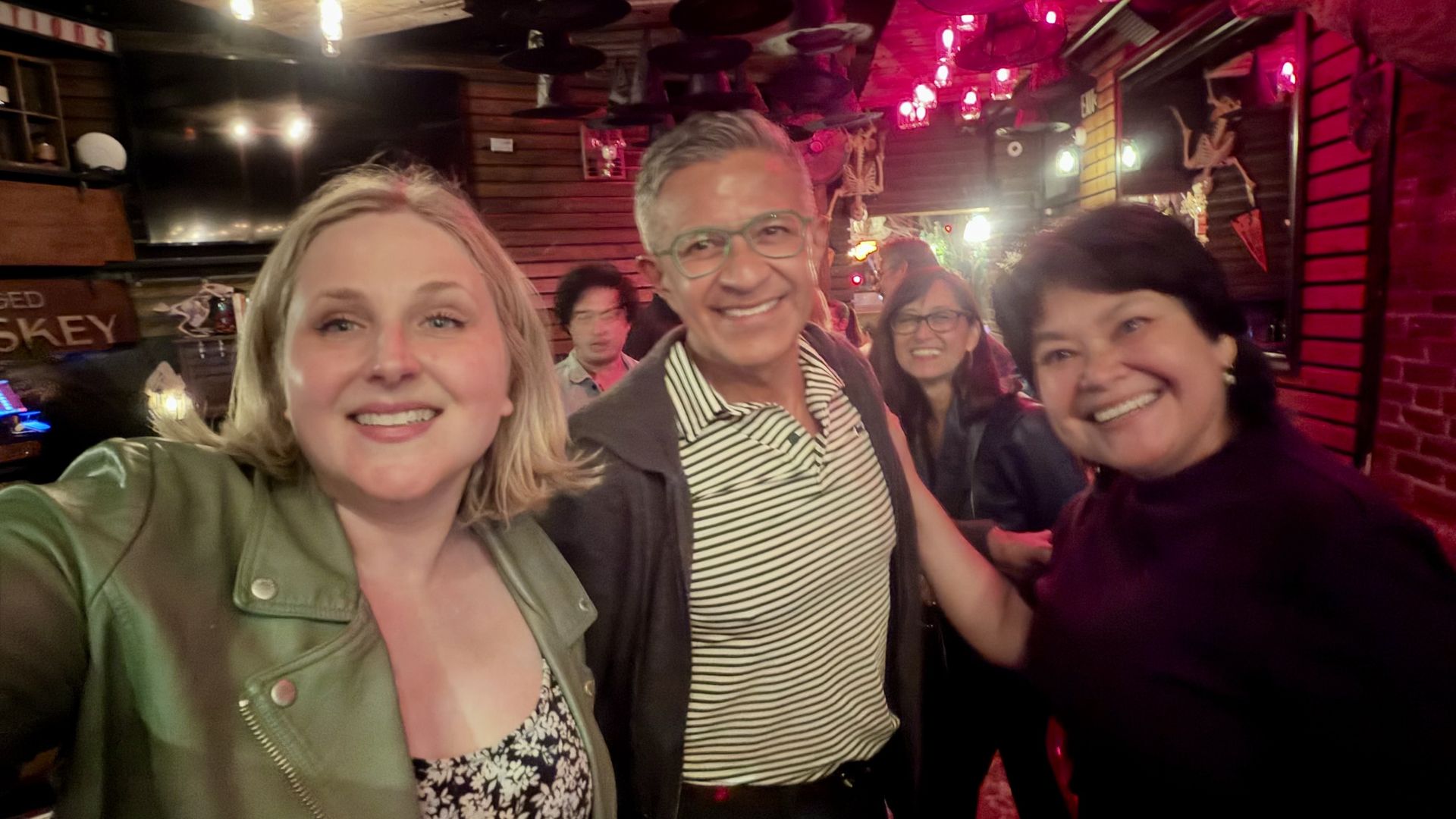 Selfie in a dim, pink-lit bar with skeleton decor and hanging hats. In the foreground, a blonde woman in a green jacket, a man with striped shirt and glasses, and a smiling woman on the right.