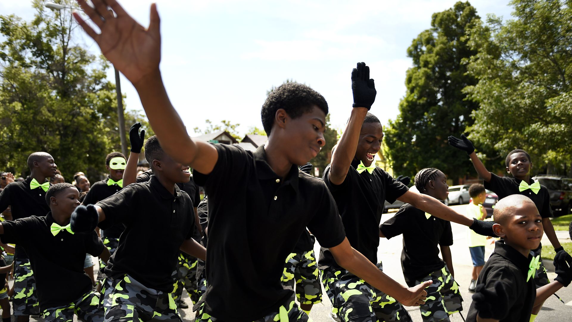 Young dancers in black shirts and neon-green camouflage shorts perform a street parade, many with gloves and neon-green bow ties, raising their arms under leafy trees.