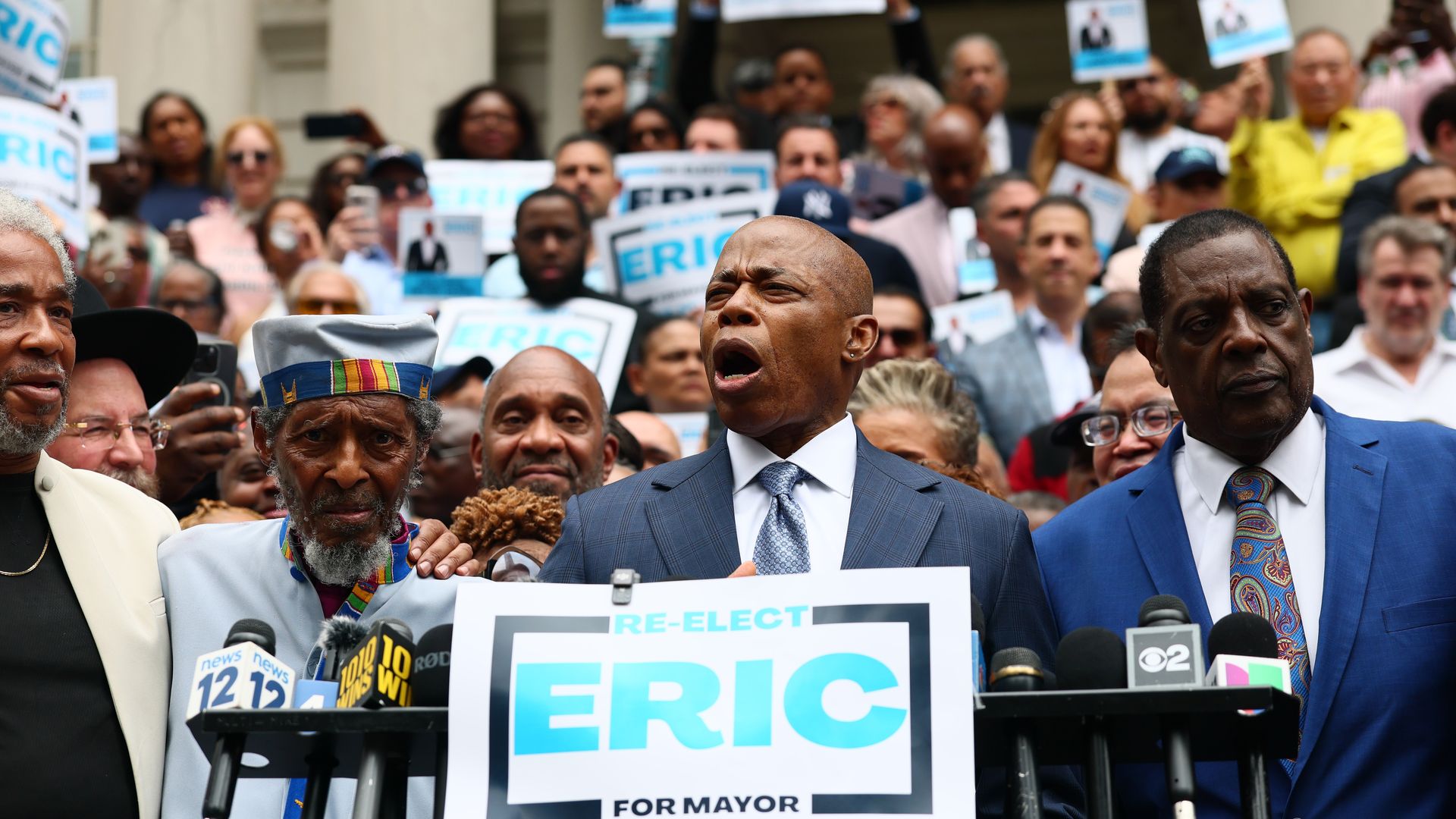 Eric Adams speaks at a campaign event behind a sign that says "RE-ELECT ERIC FOR MAYOR" while supporters stand behind him.
