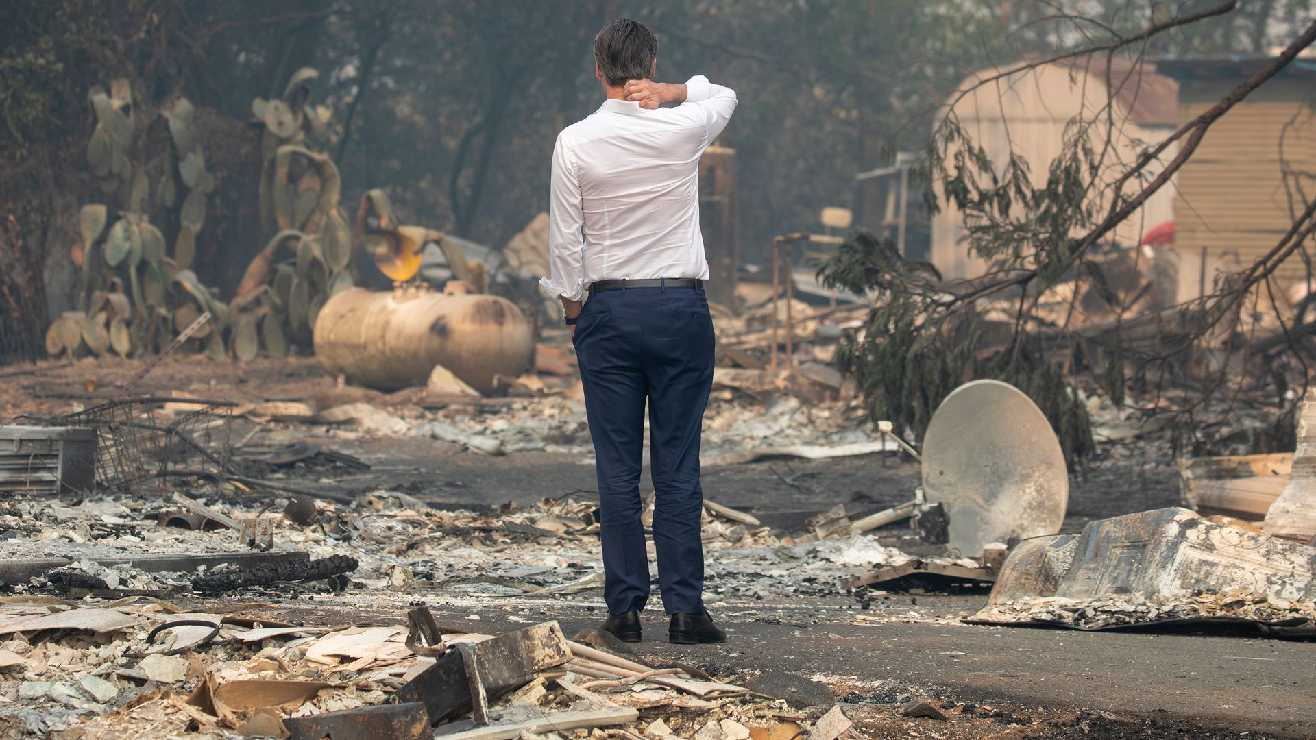In this image, Gavin Newsom stands in a dress shirt and slacks in the middle of a rubble-filled, burned-out street after a large fire