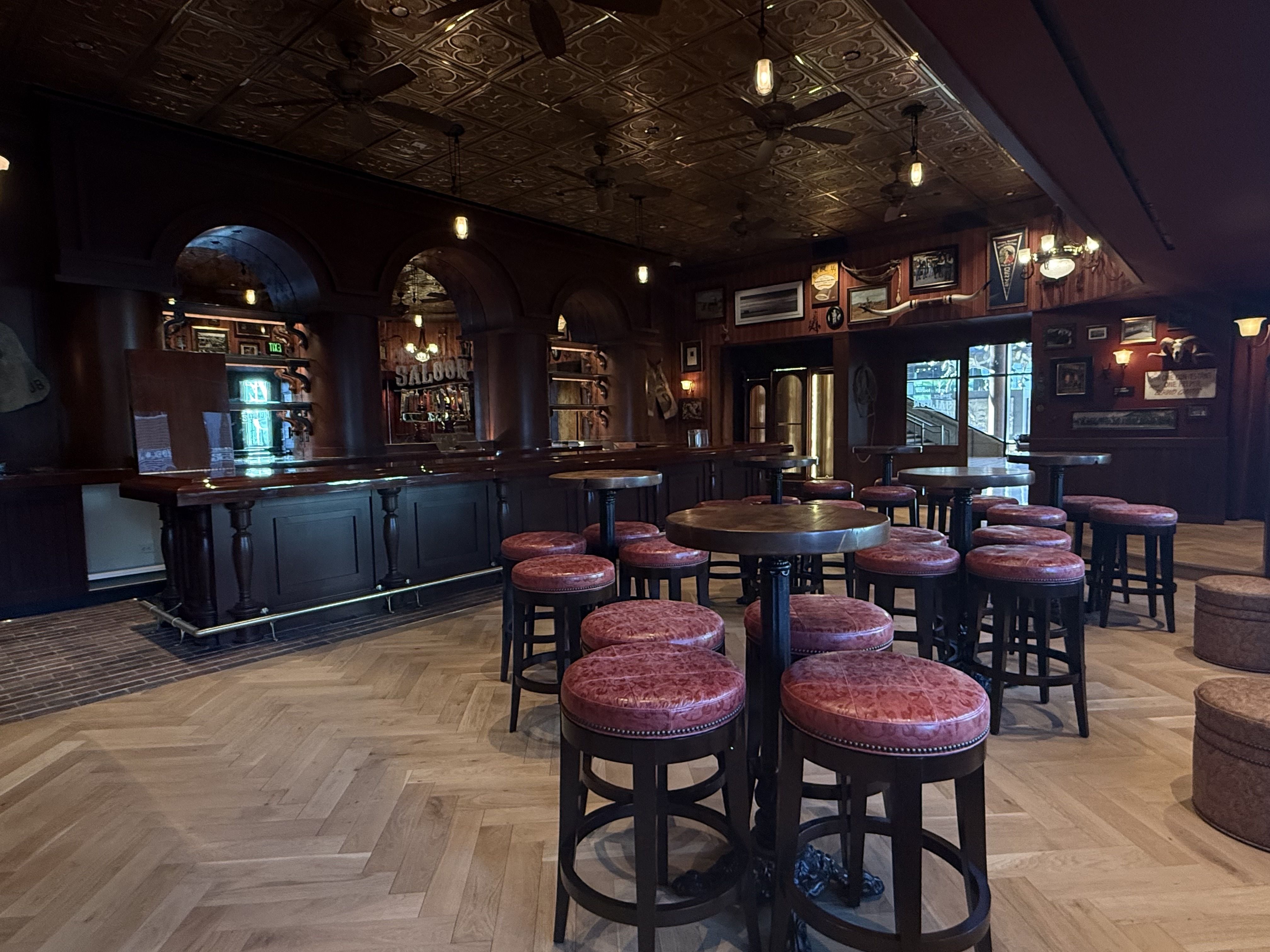 Interior of a dimly lit, vintage-style wooden bar with round tables and red upholstered stools, framed pictures and a sign reading "Saloon" on the back wall.