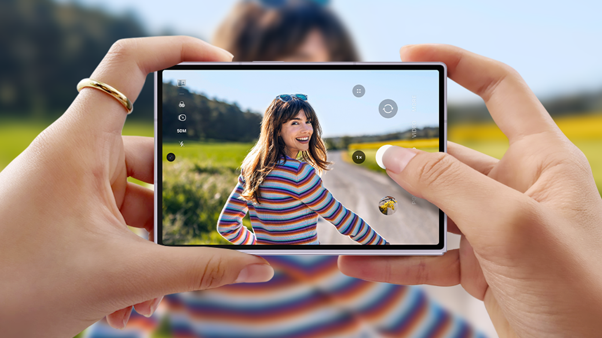 Two hands hold a smartphone to photograph a smiling woman in a bright striped top outdoors on a sunny day. The phone's screen shows the scene with a road, green fields, and blue sky.