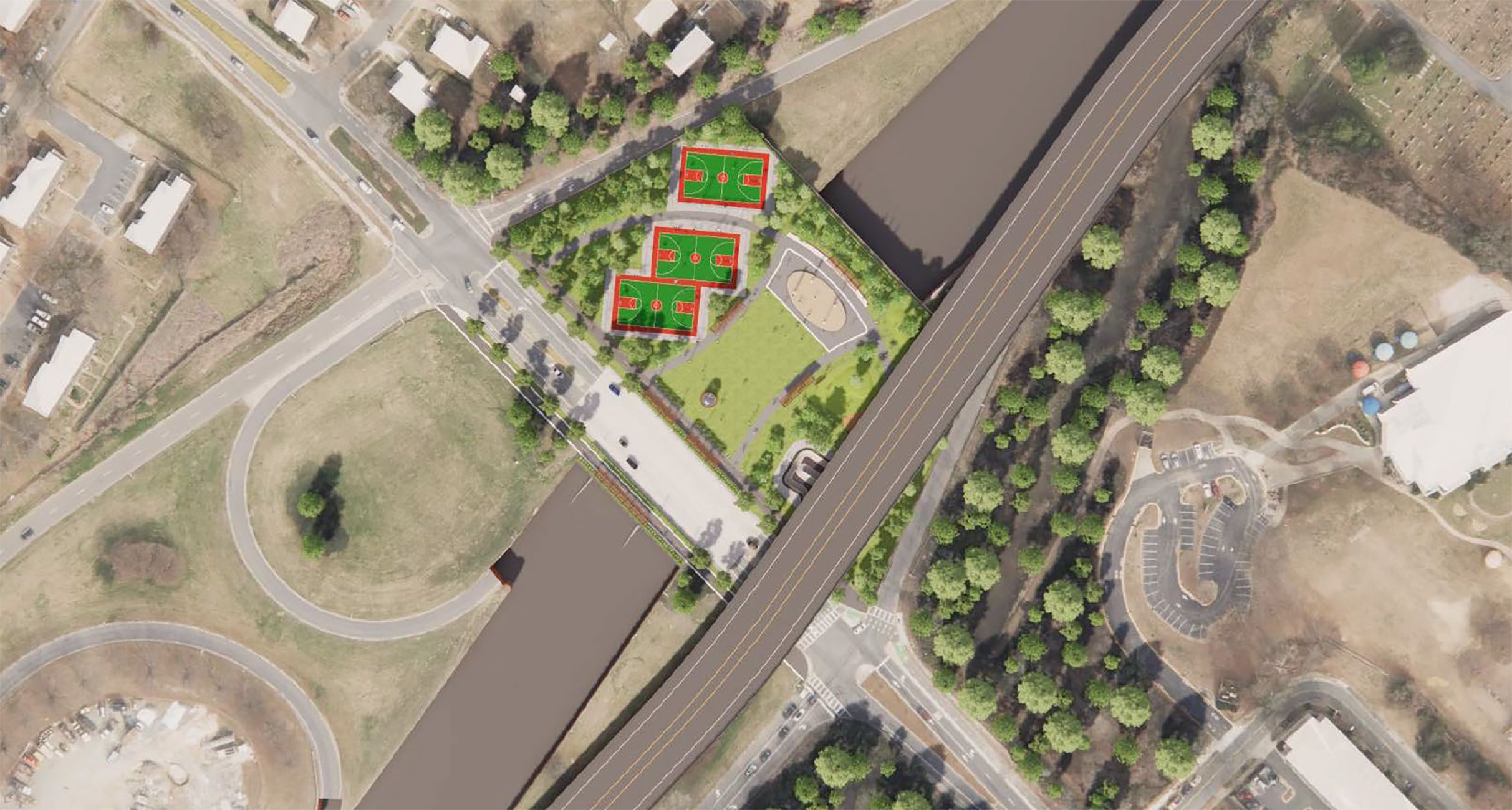 An aerial view of a park showing three green courts with red borders beside a river, near a highway. Trees line walkways and a parking lot sits nearby.