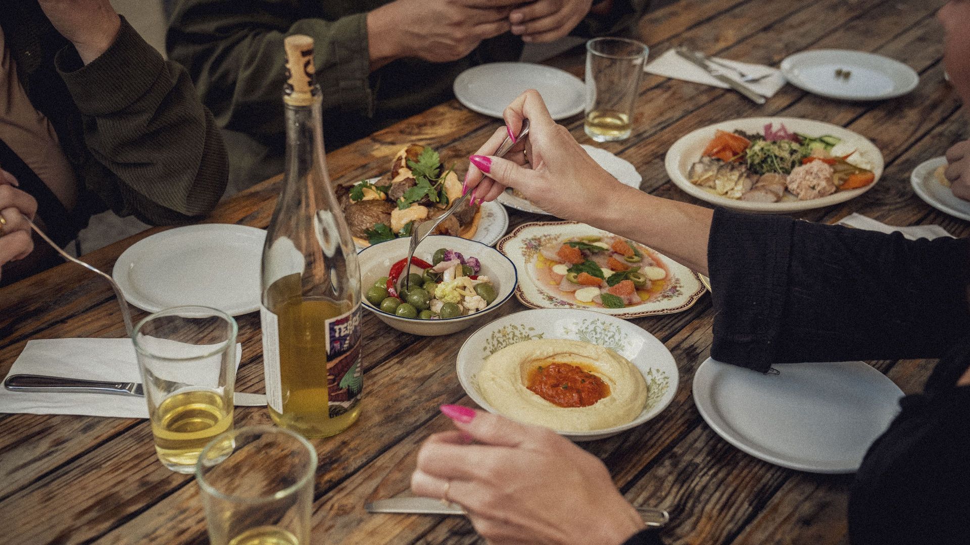 People share assorted small plates like hummus, olives, fish and wine on an outdoor patio table. 
